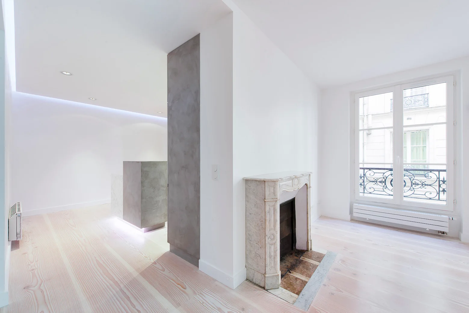 An empty minimalist white room featuring light wood flooring, a grey concrete column, and a traditional marble fireplace.