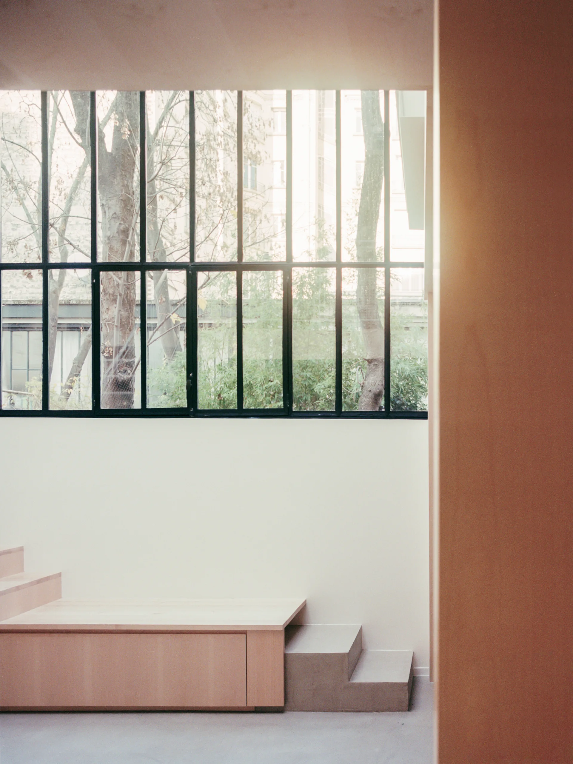 A light wood tiered platform bench sits against a plain off-white wall below a large black metal grid window showing outdoor trees.