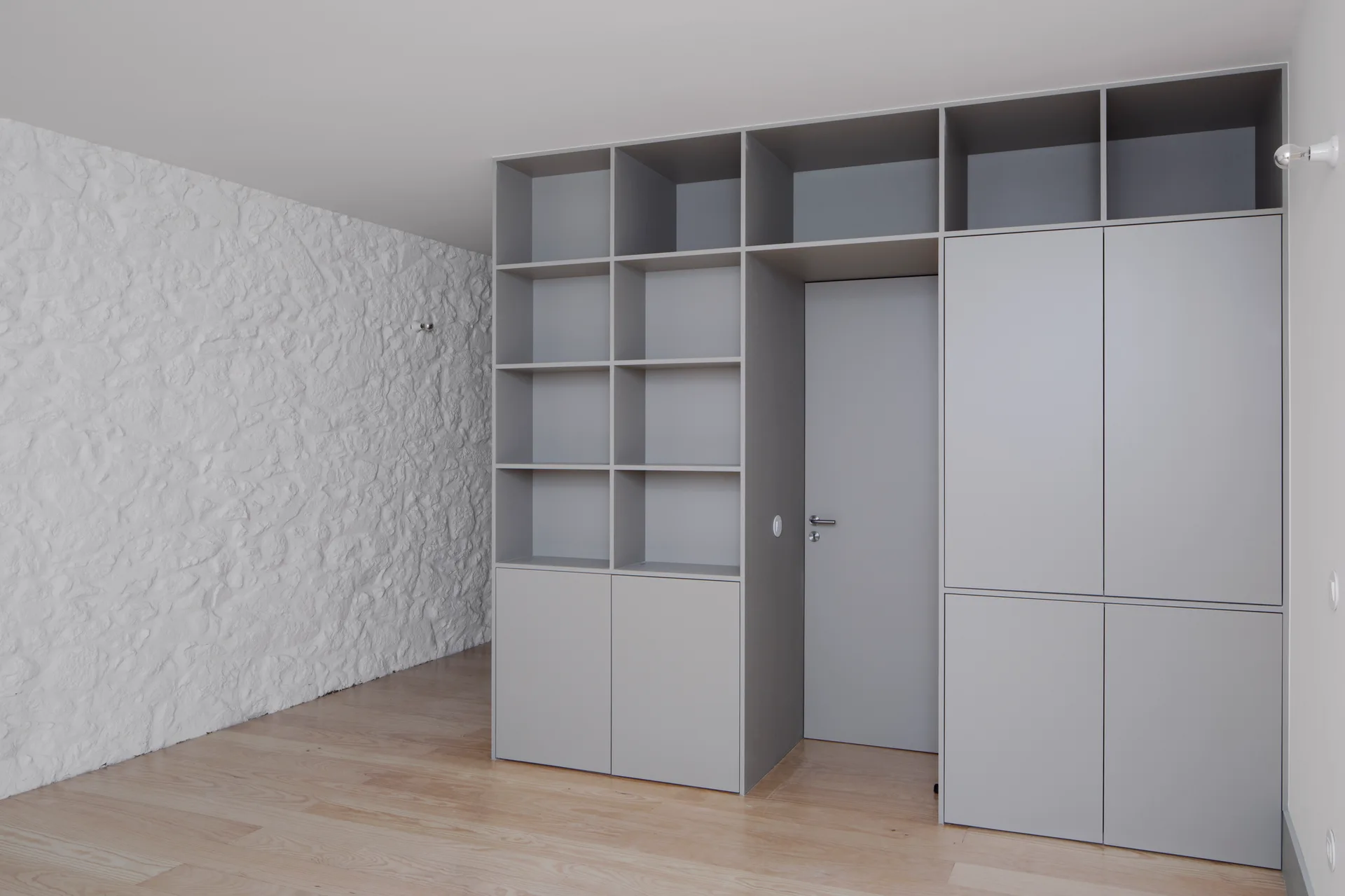 A custom floor-to-ceiling grey wardrobe unit integrates a matching door next to a white textured stone wall and light wood flooring.
