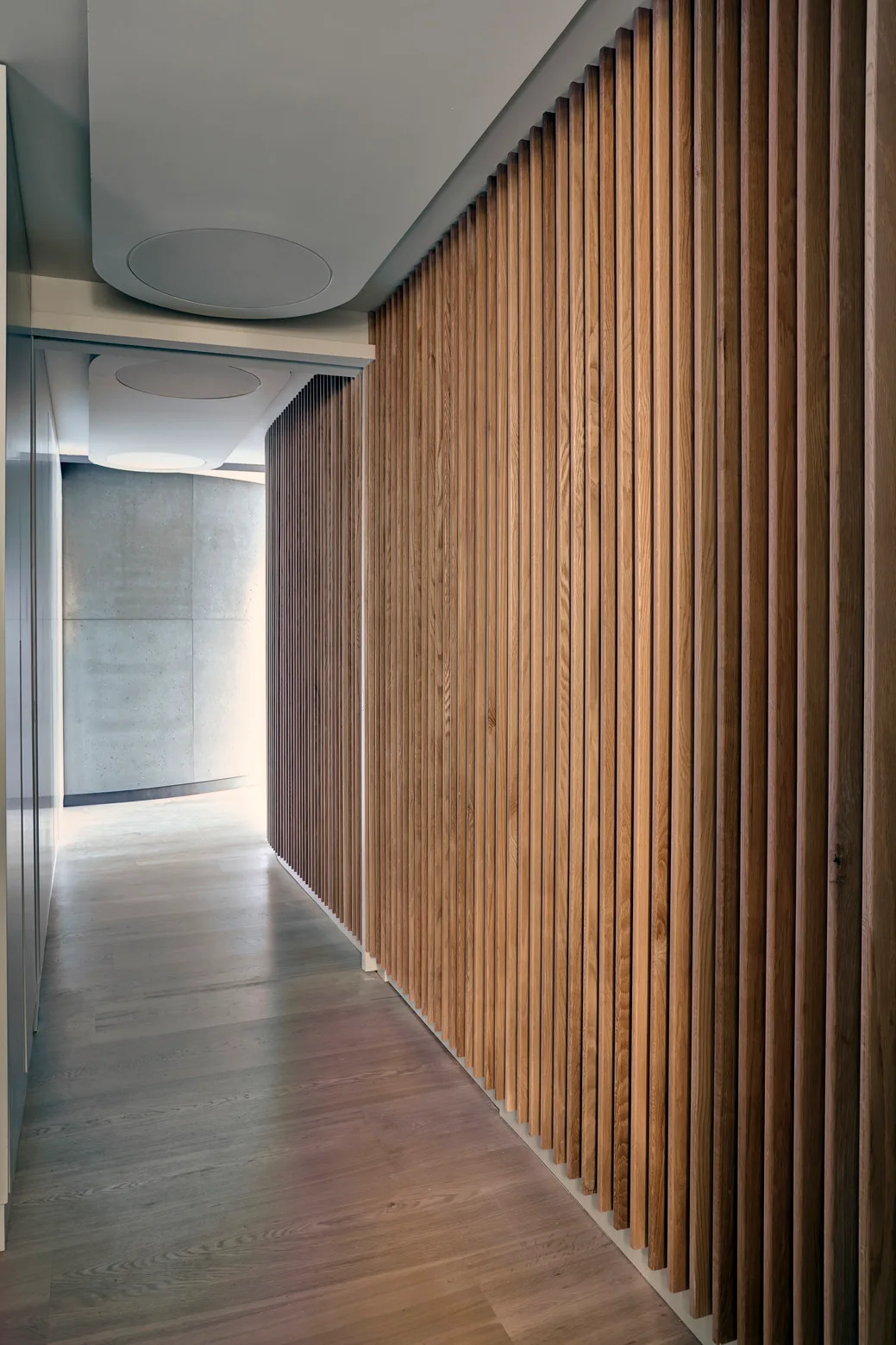 A modern hallway featuring a vertical wood slatted accent wall, light wood flooring, and a smooth concrete wall at the far end.