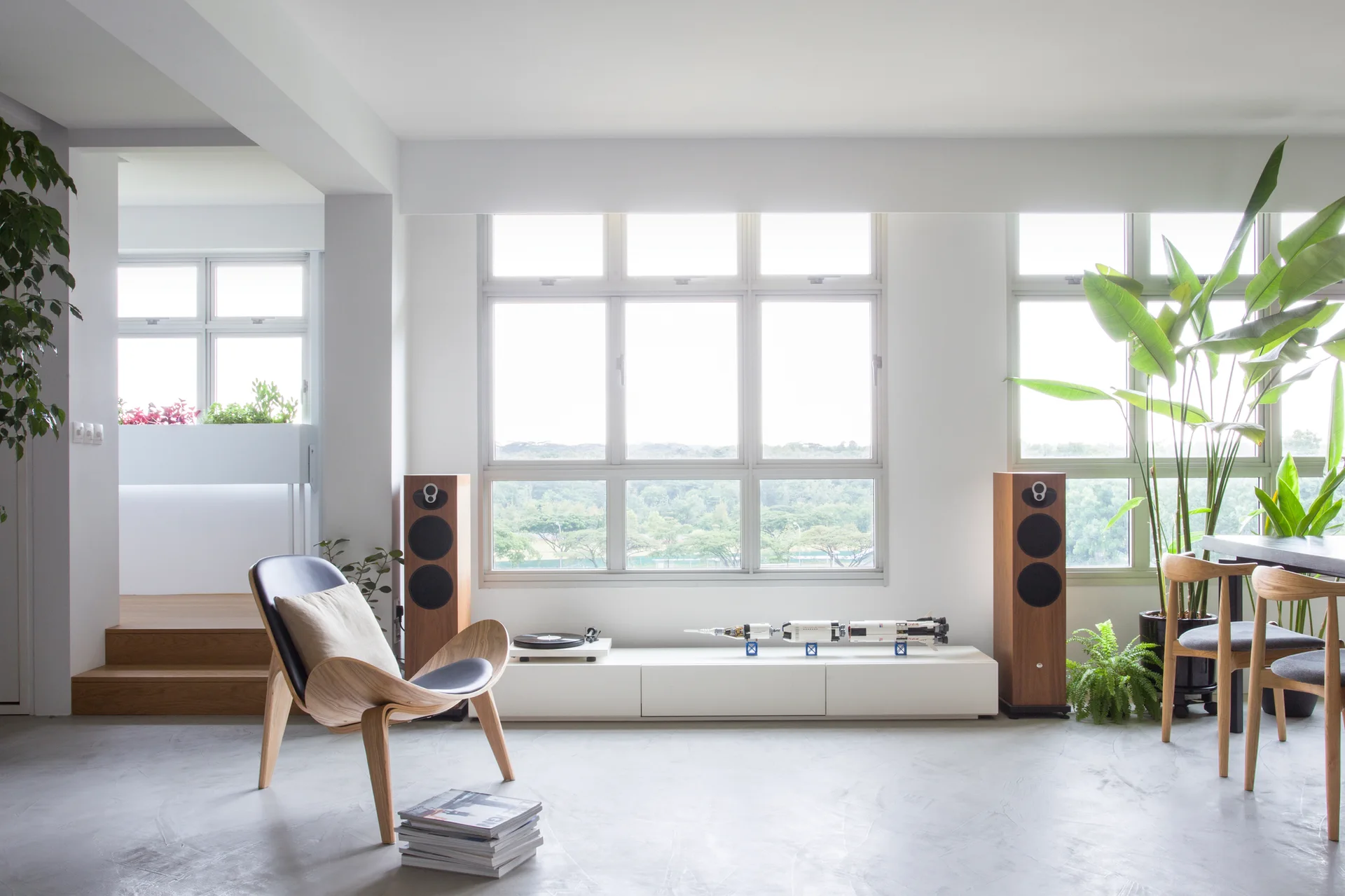 A modern living room features a curved wood lounge chair, concrete floors, and a low white media console flanked by tall wooden speakers.