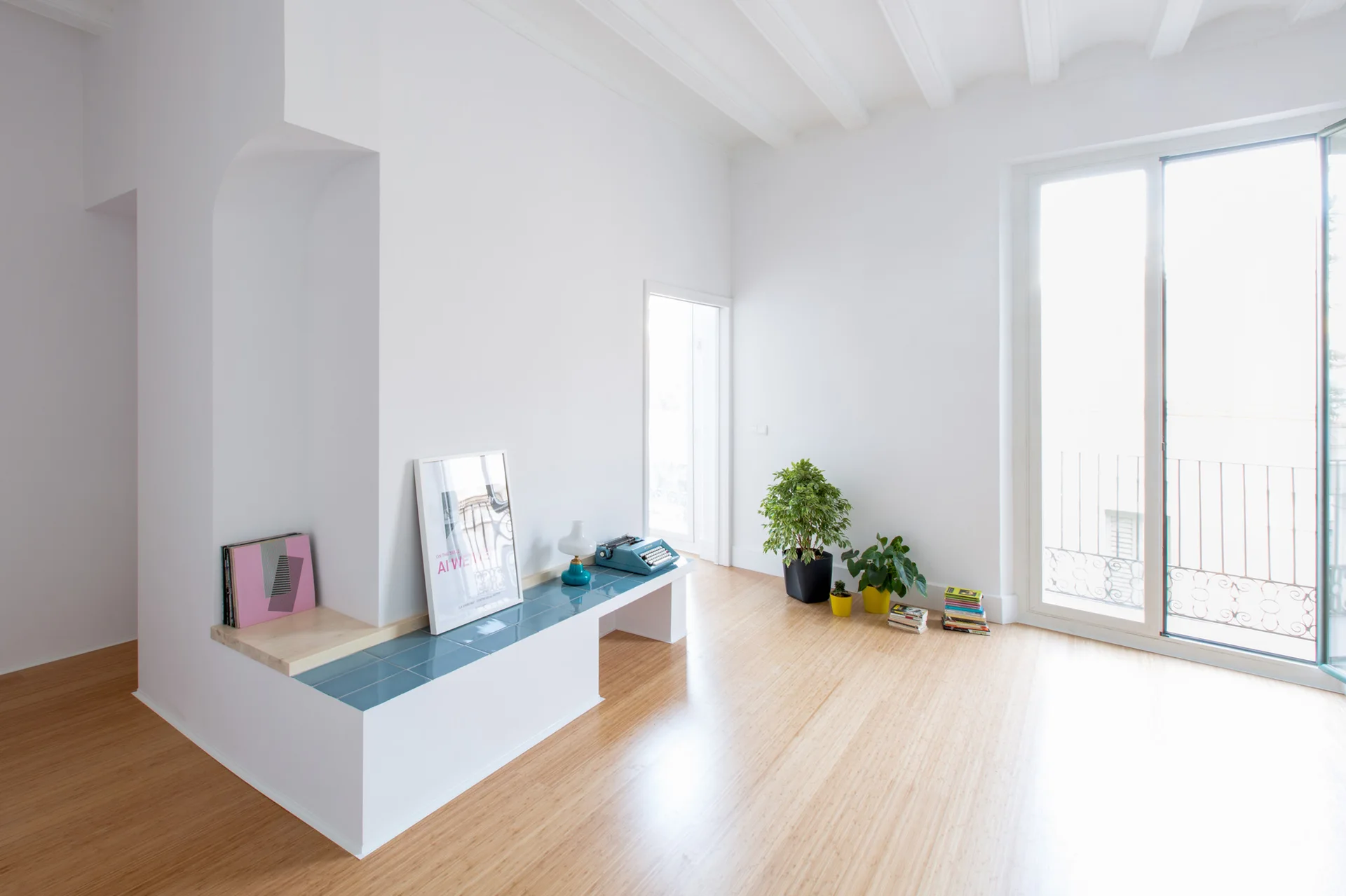 A minimalist white room with light wood floors, an arched wall niche, and a low built-in bench topped with glossy blue tiles.