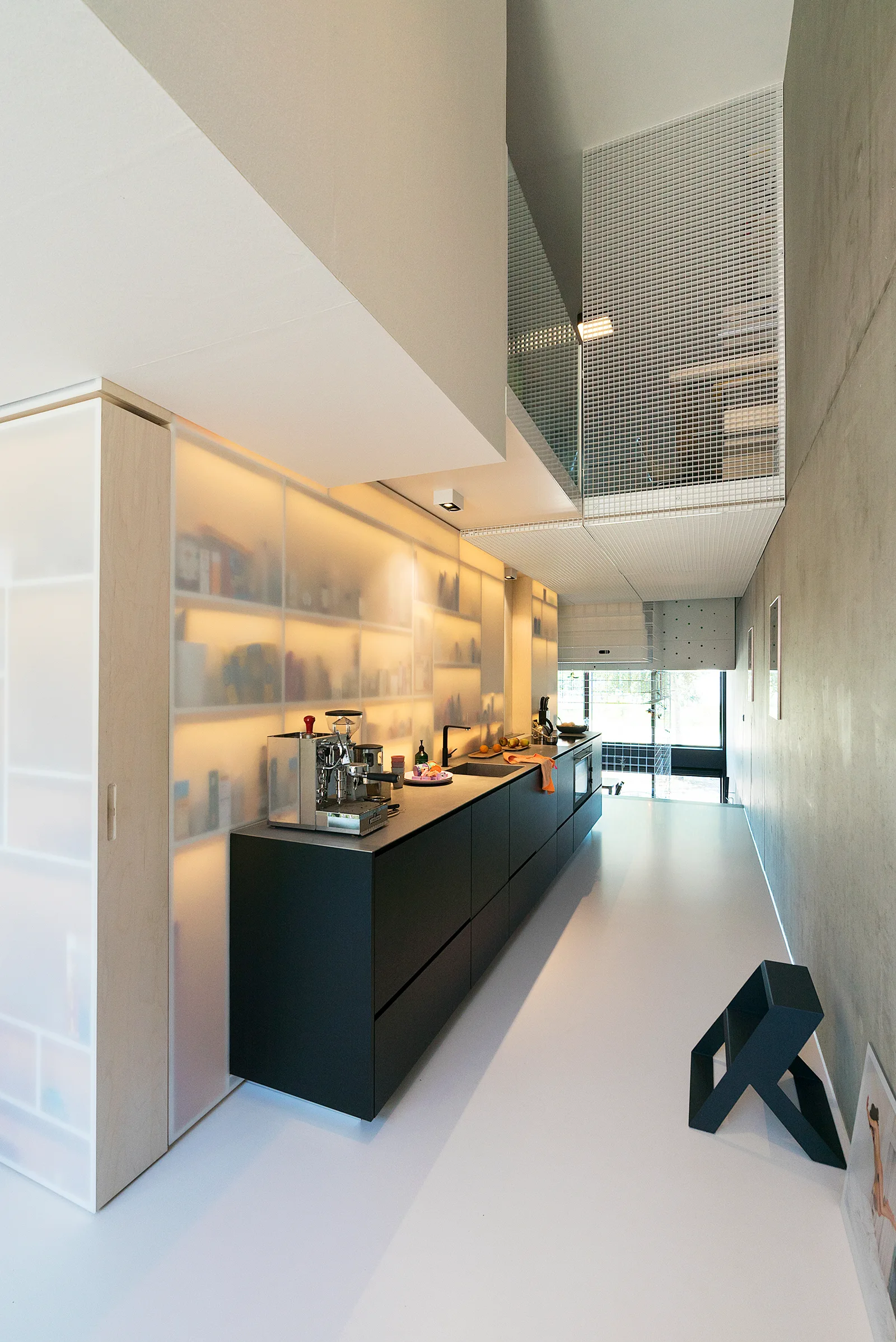 A narrow minimalist kitchen features black base cabinets beneath a backlit frosted glass storage wall, a concrete wall, and a seamless white floor.