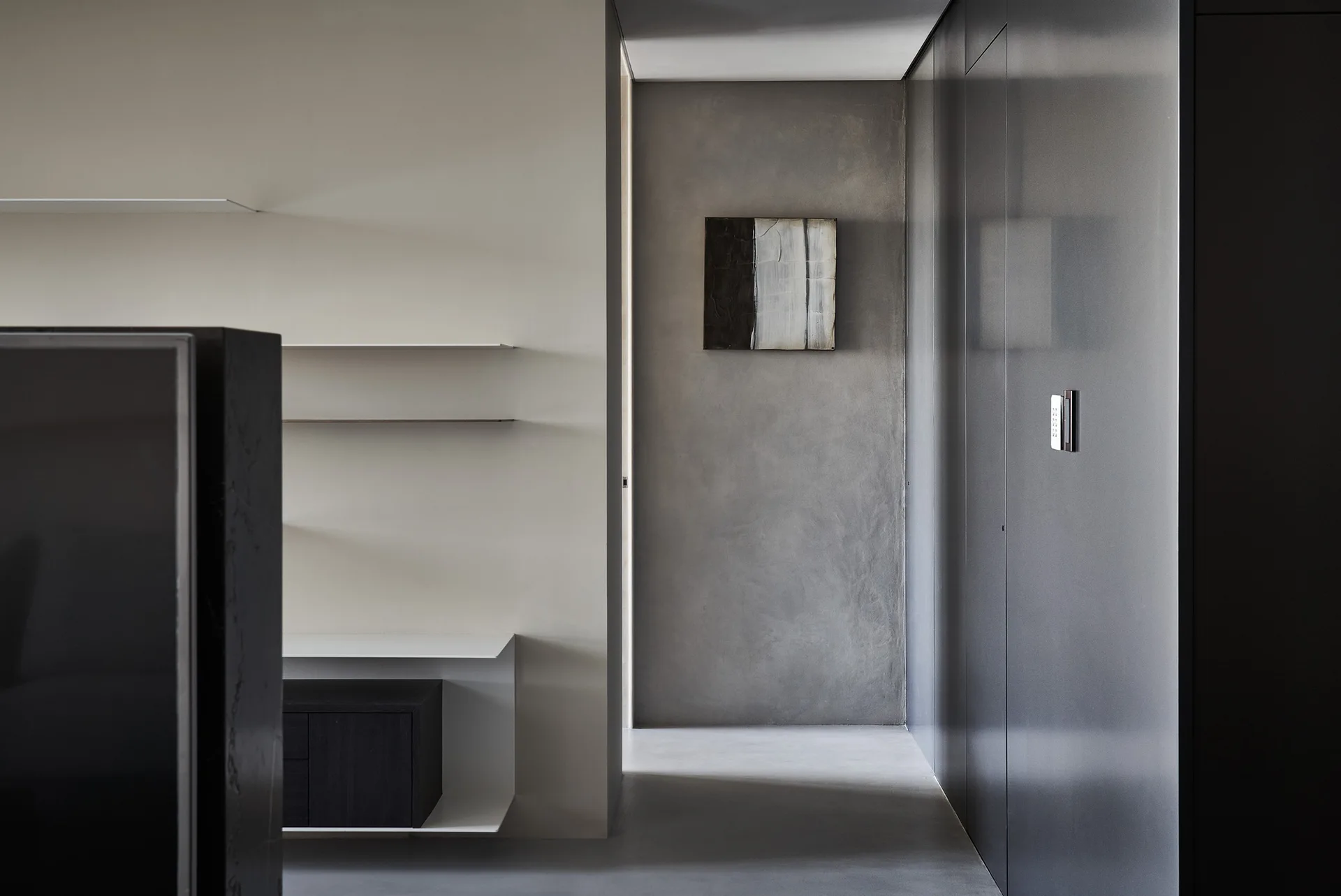 A minimalist hallway features built-in white shelving, dark wall paneling, and a textured grey plaster wall decorated with a small abstract artwork.