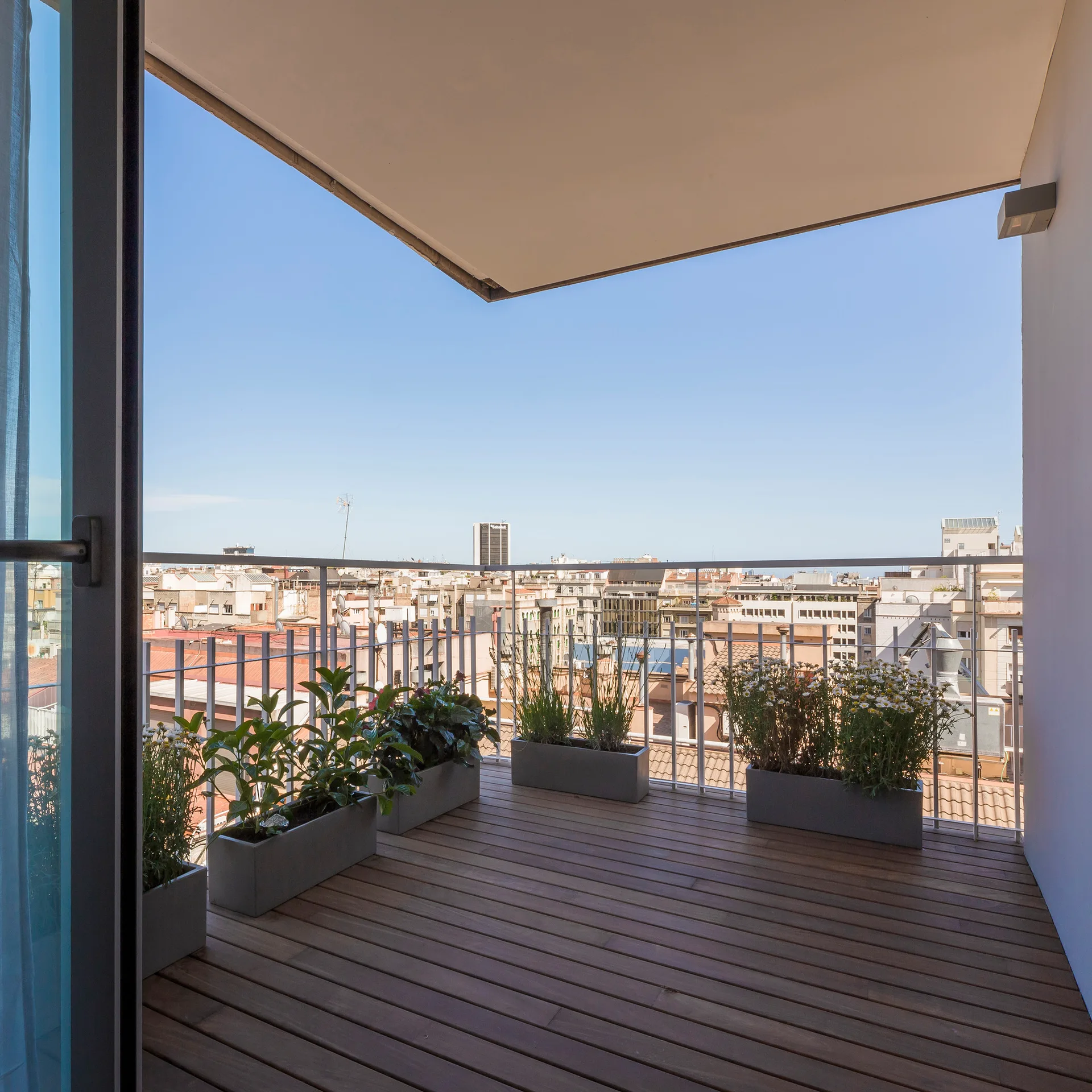 A modern balcony with dark wood plank flooring, several low grey planters with green foliage, and a thin metal railing overlooking a cityscape.