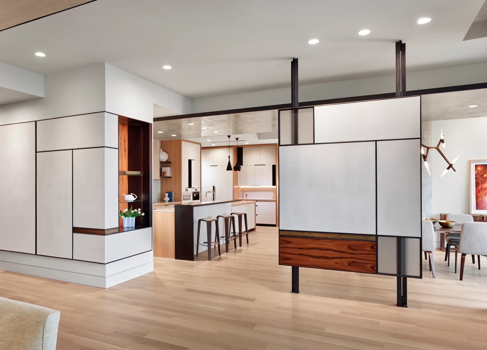 A geometric room divider with black framing and wood panels separates a light-filled hallway from an open kitchen and dining area.