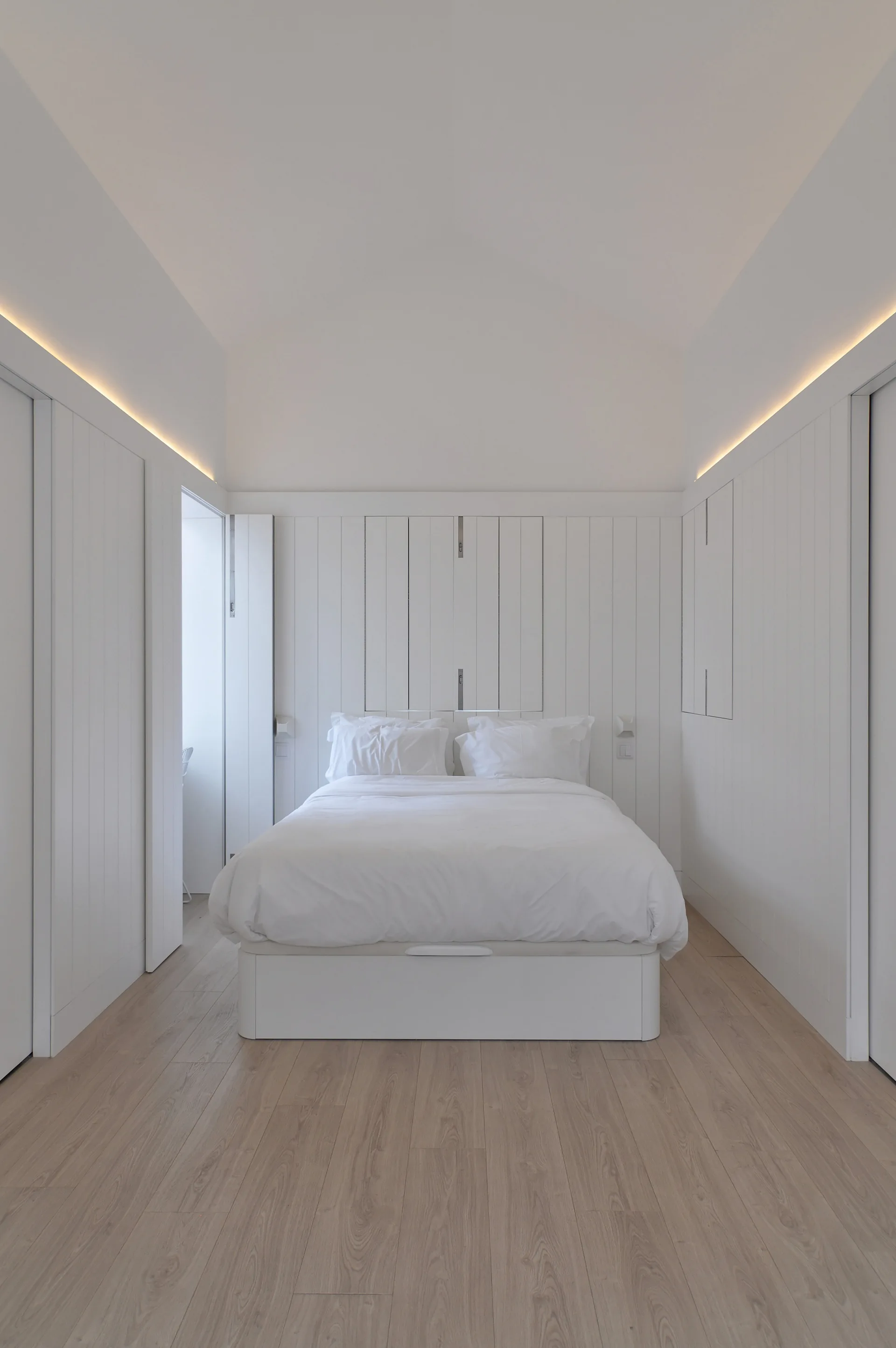 Minimalist white bedroom featuring vertical wall paneling, built-in storage, light wood flooring, and warm cove lighting above a white bed.