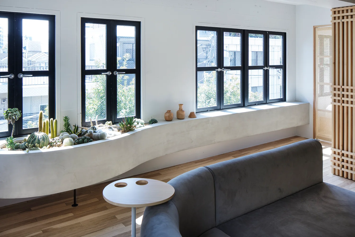 A bright living room features a long curved concrete planter filled with cacti, situated beneath black-framed windows next to a gray sofa.