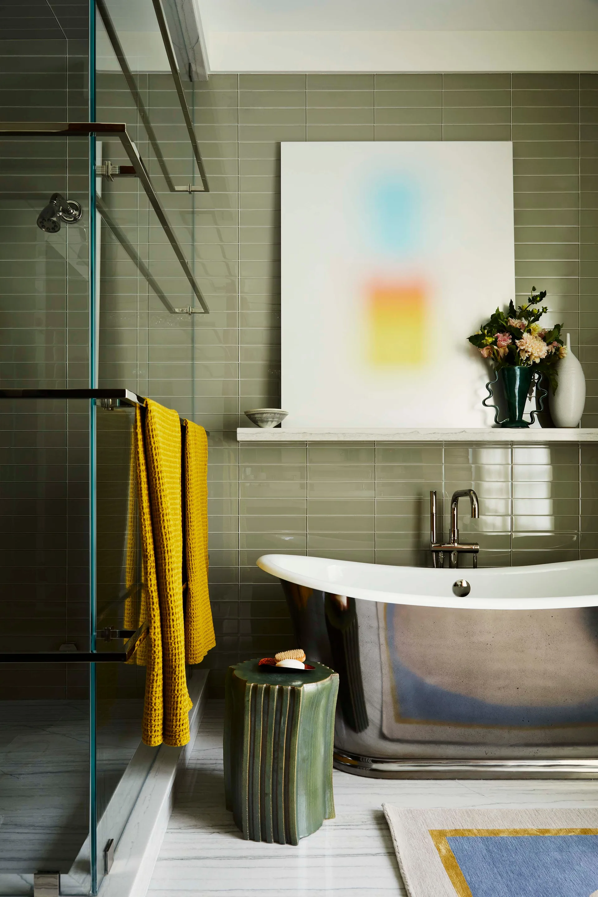 A contemporary bathroom featuring a polished metallic bathtub next to a glass shower and a sage green tiled wall with a decorative shelf.