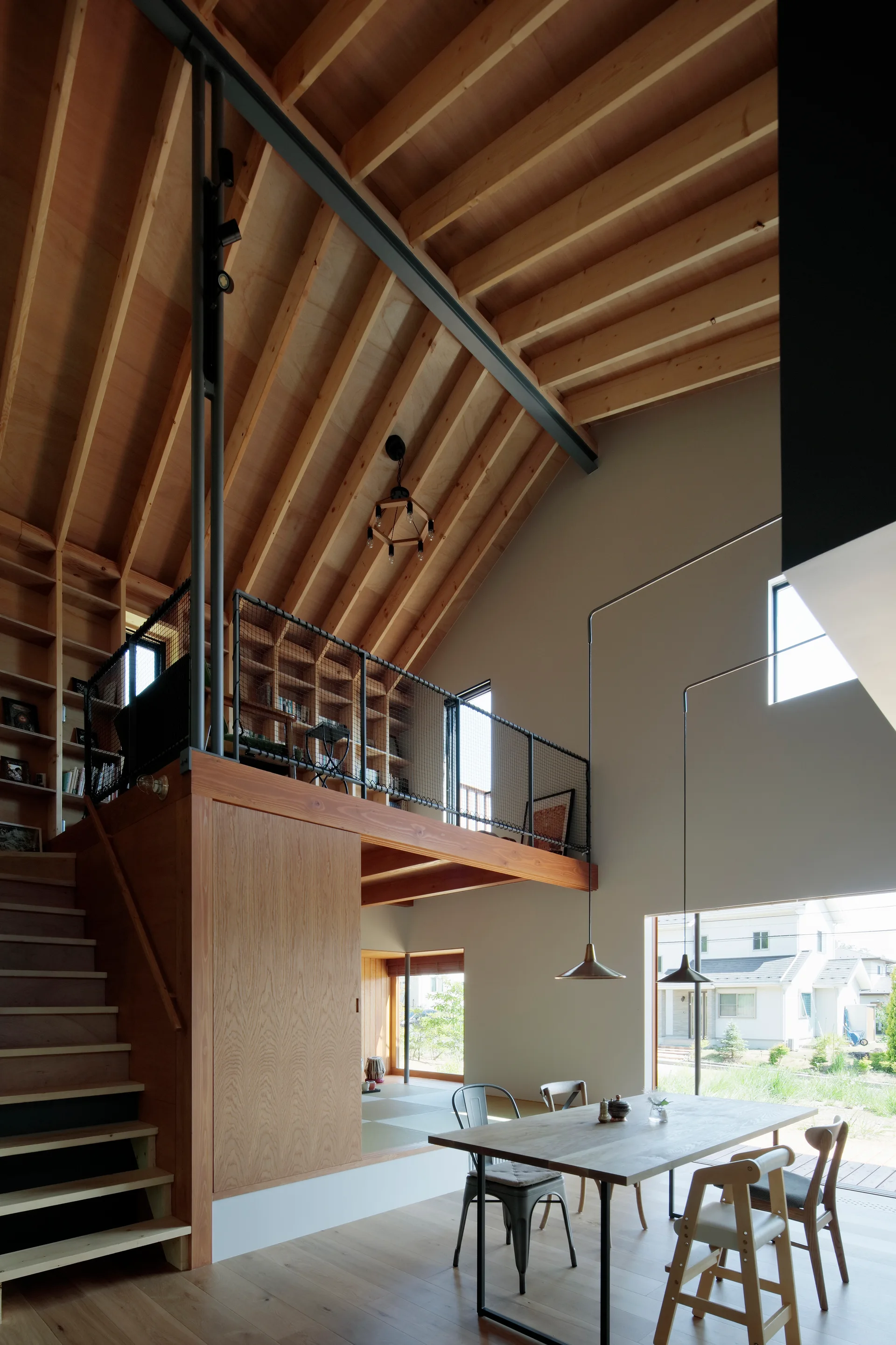 A double-height dining room featuring a wooden mezzanine loft with black mesh railings above a wood dining table with pendant lights.