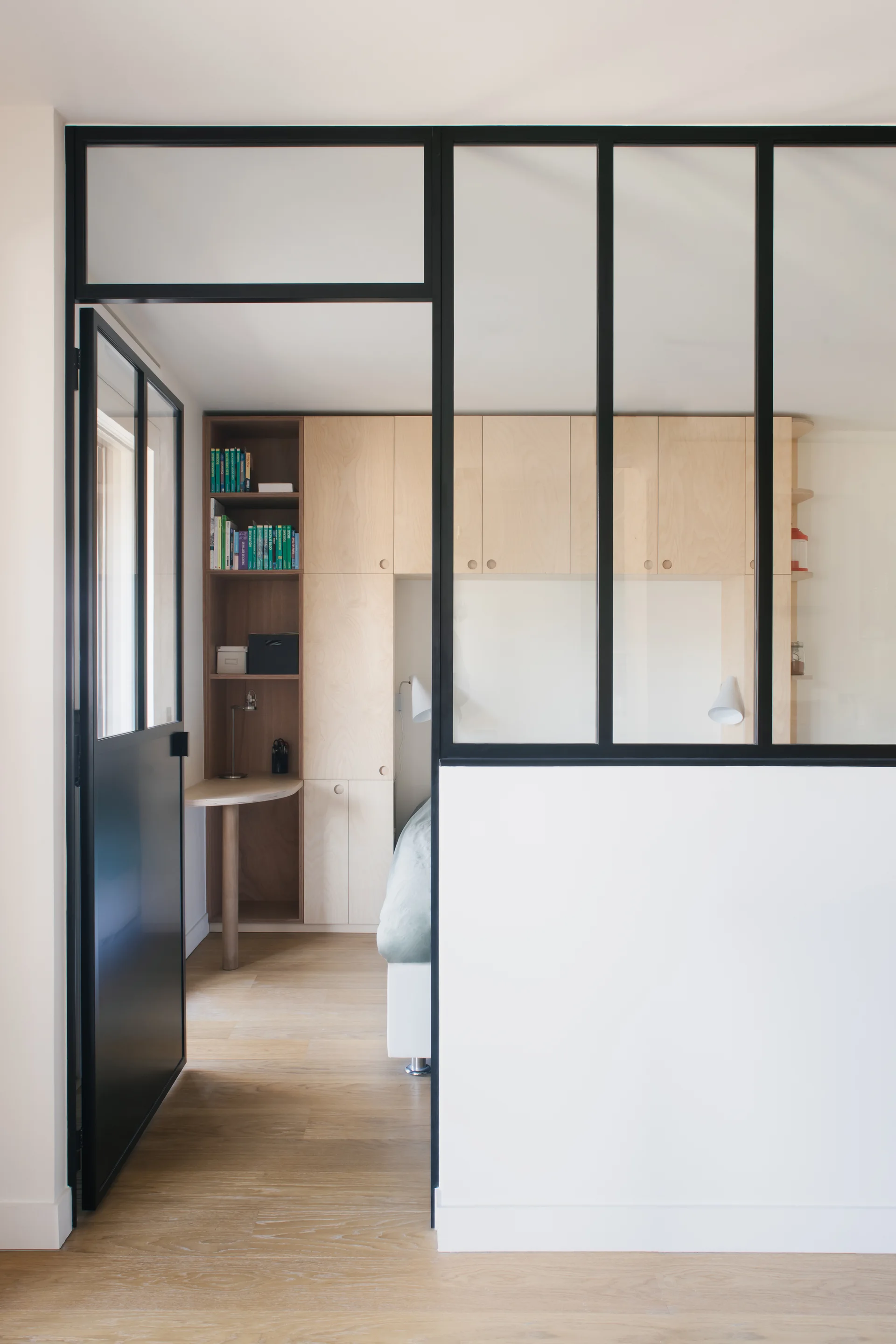 A black-framed glass partition wall opens to a bedroom featuring custom light wood storage cabinets, a built-in desk, and a bed.