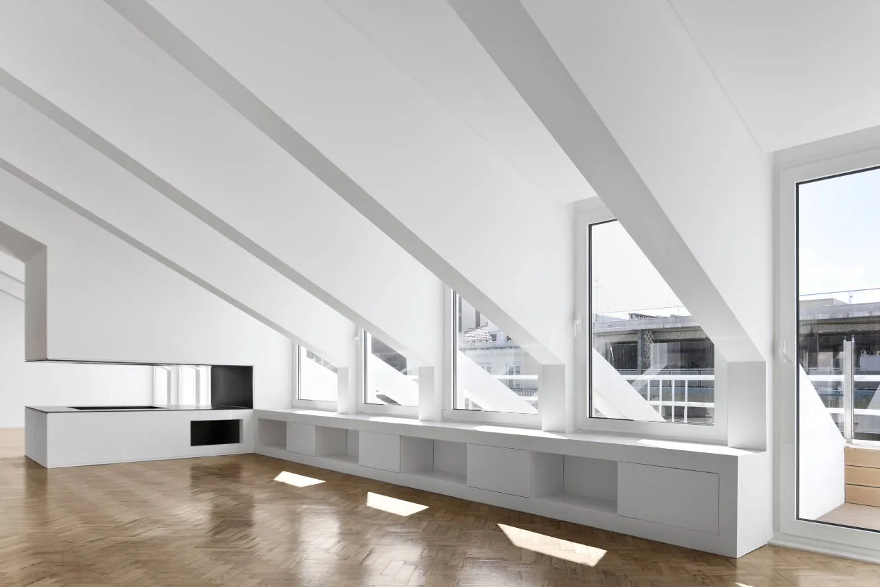 An empty attic room with a white sloped ceiling, herringbone wood parquet flooring, and a long low built-in storage cabinet under large windows.