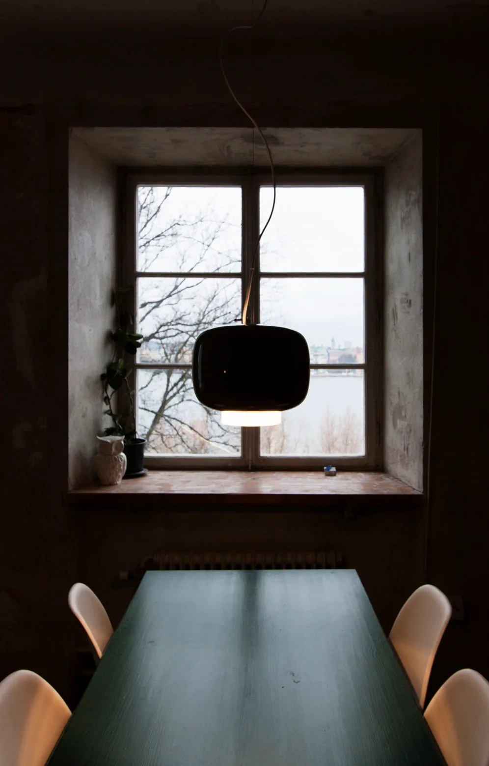 A dark green dining table with white chairs sits beneath a black dome pendant light in front of a window overlooking a body of water.