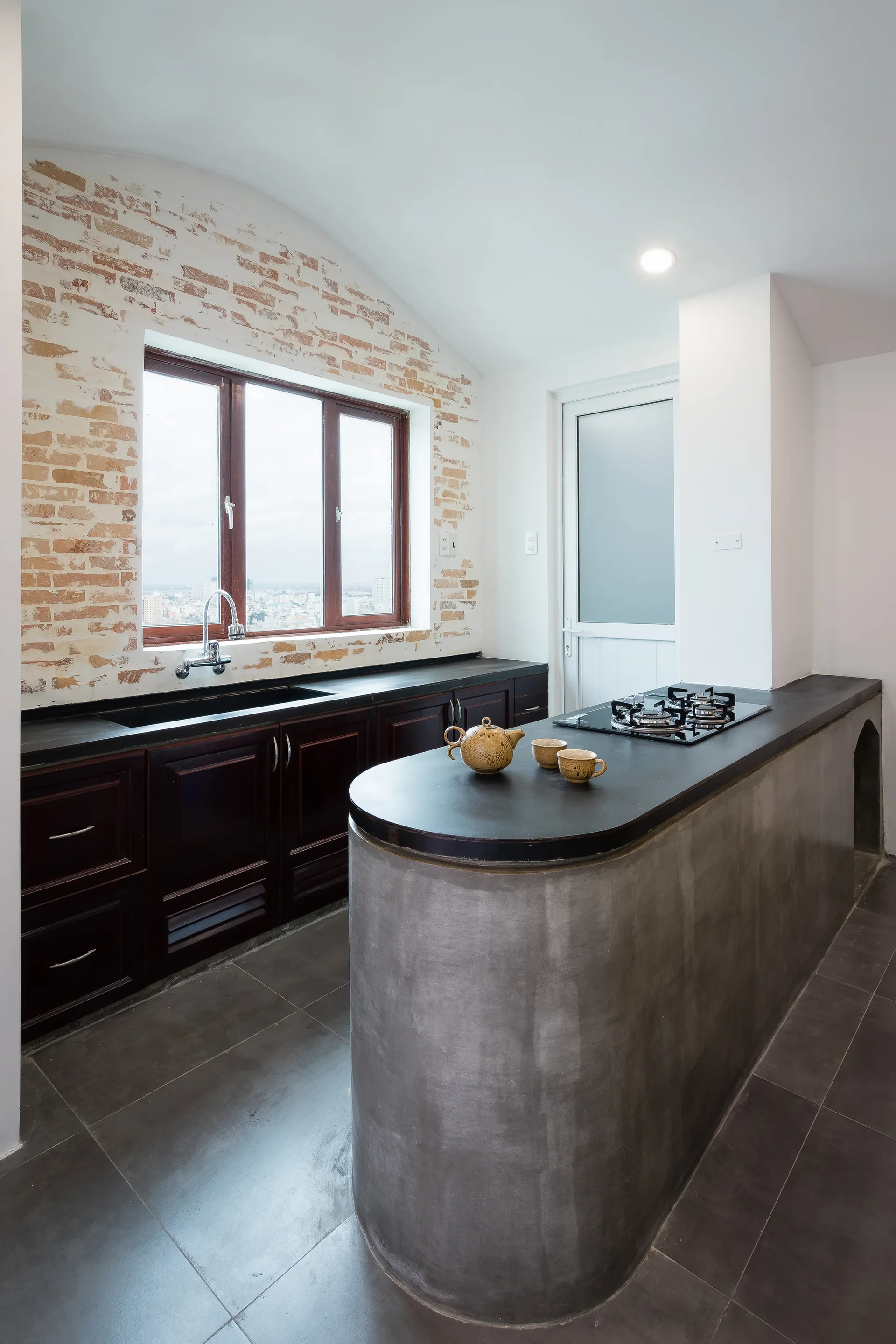 An industrial kitchen featuring a curved concrete island with a gas cooktop, dark wood cabinets, and an exposed brick wall around a large window.