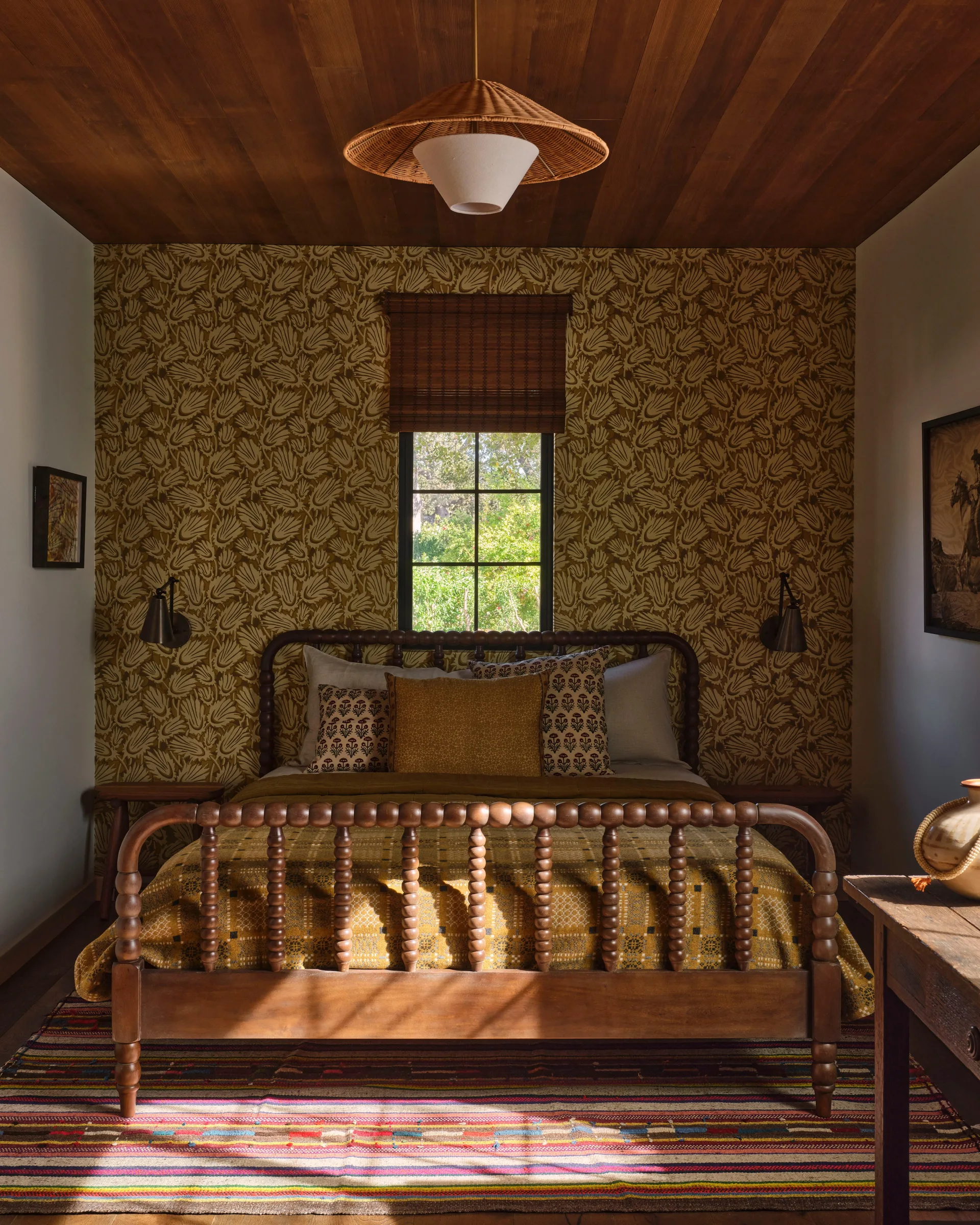 A wooden spindle bed with mustard yellow bedding is positioned against botanical patterned wallpaper under a dark wood plank ceiling.