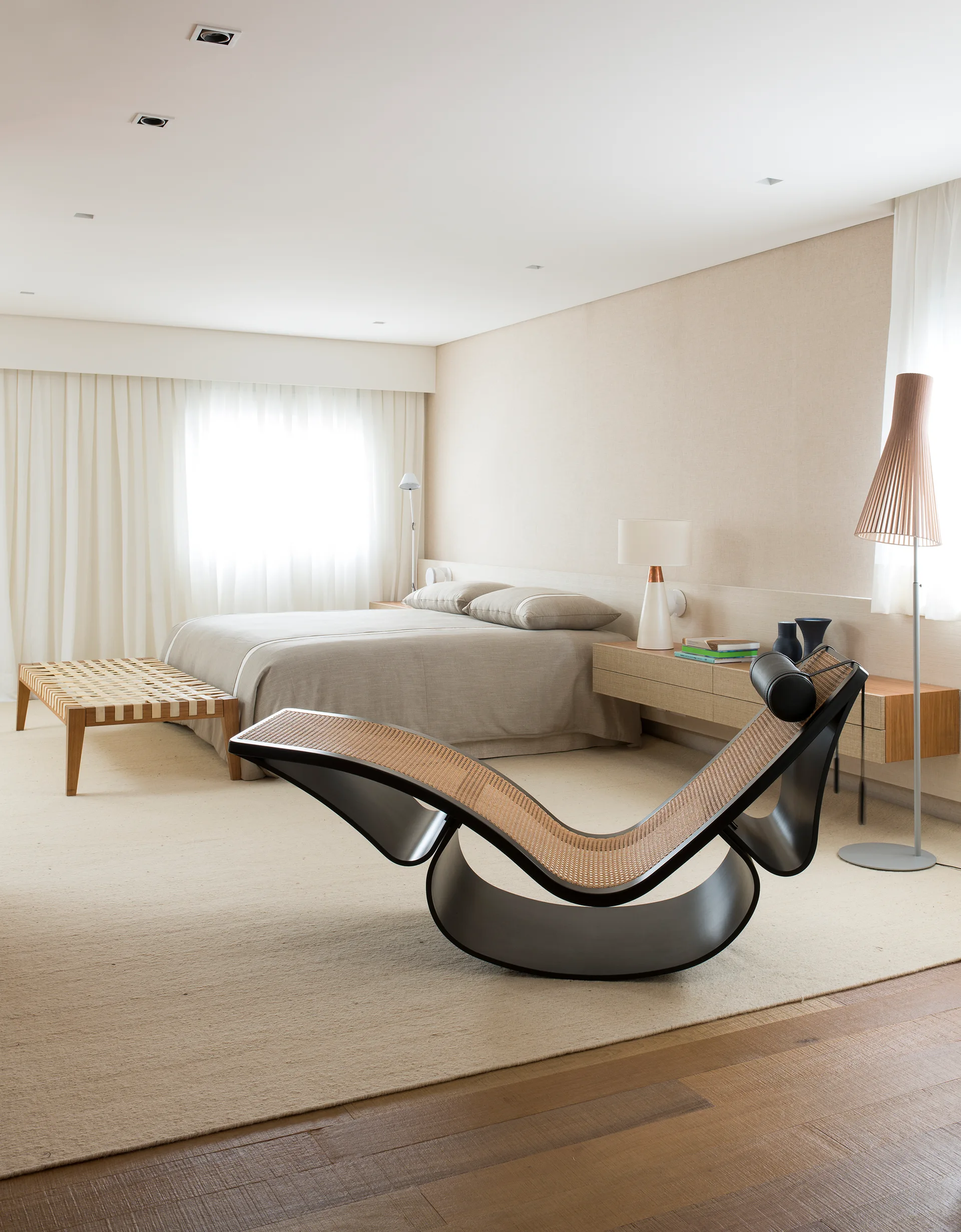 A minimalist neutral bedroom features a sculptural black curved chaise lounge, a beige bed, and wall-mounted bedside storage.