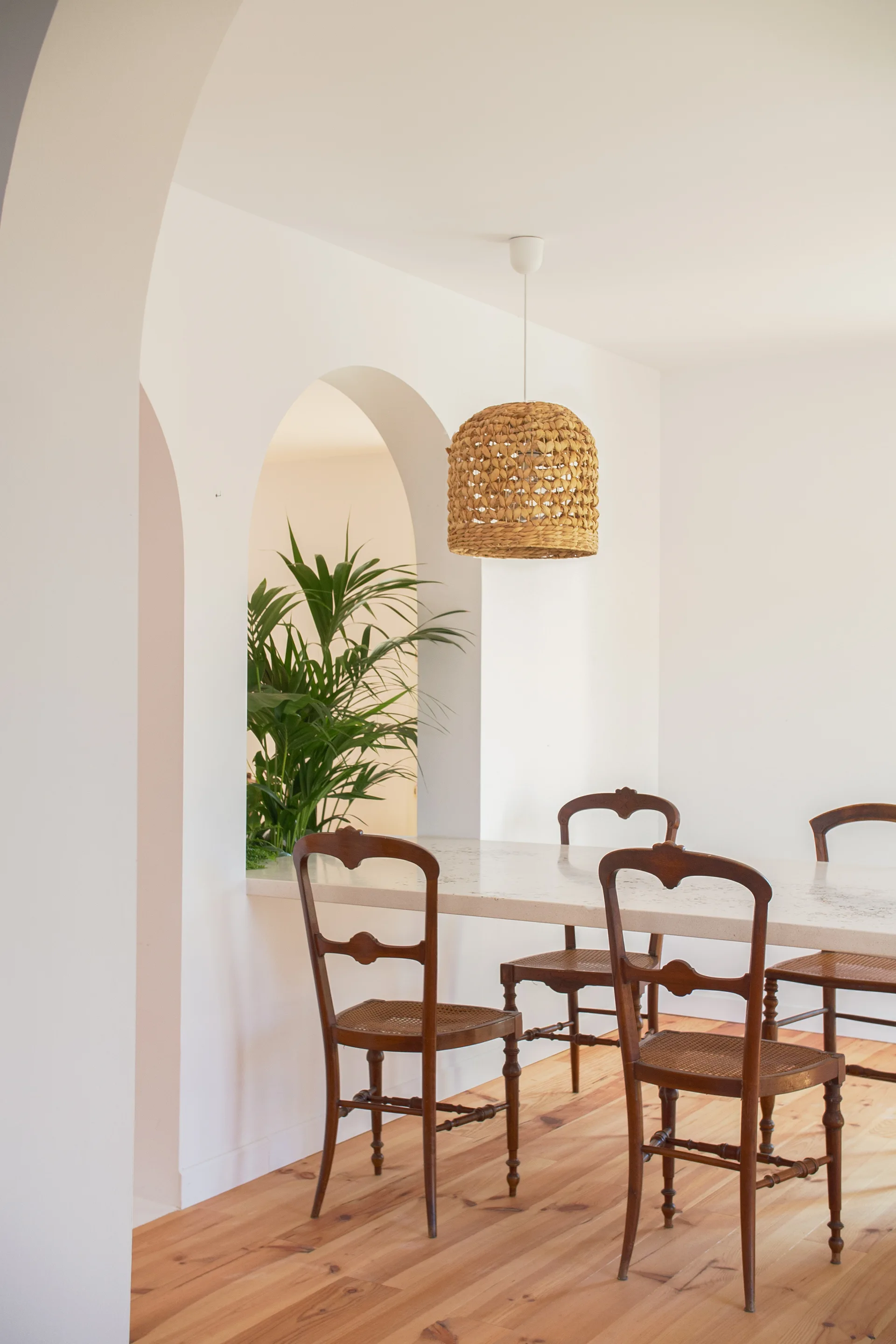 A bright dining area features antique wooden chairs around a stone table, underneath a woven pendant light next to a white plaster archway.