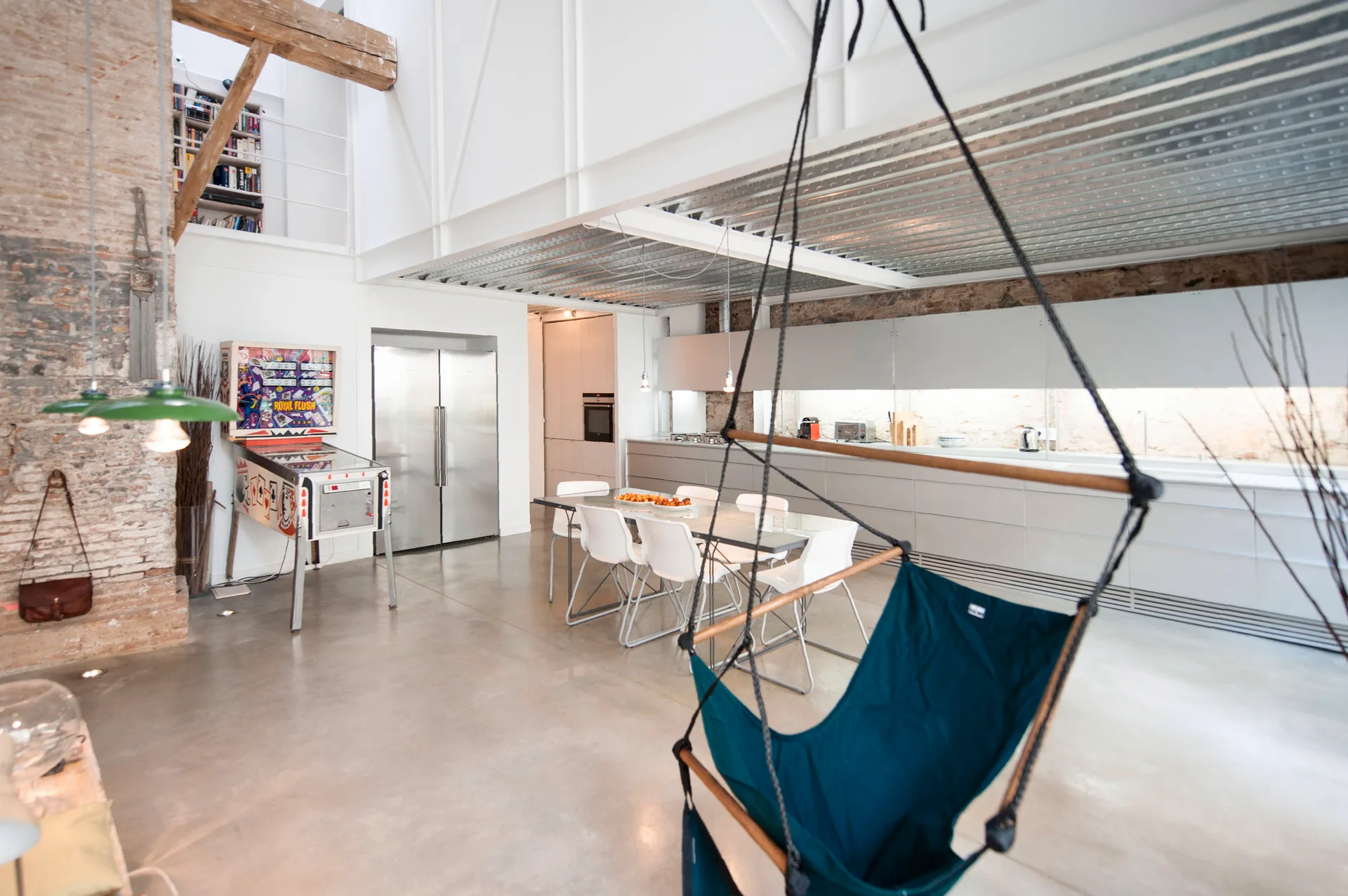 An open plan loft kitchen with concrete floors, white cabinets, a dining table, a vintage pinball machine, and a hanging swing chair.