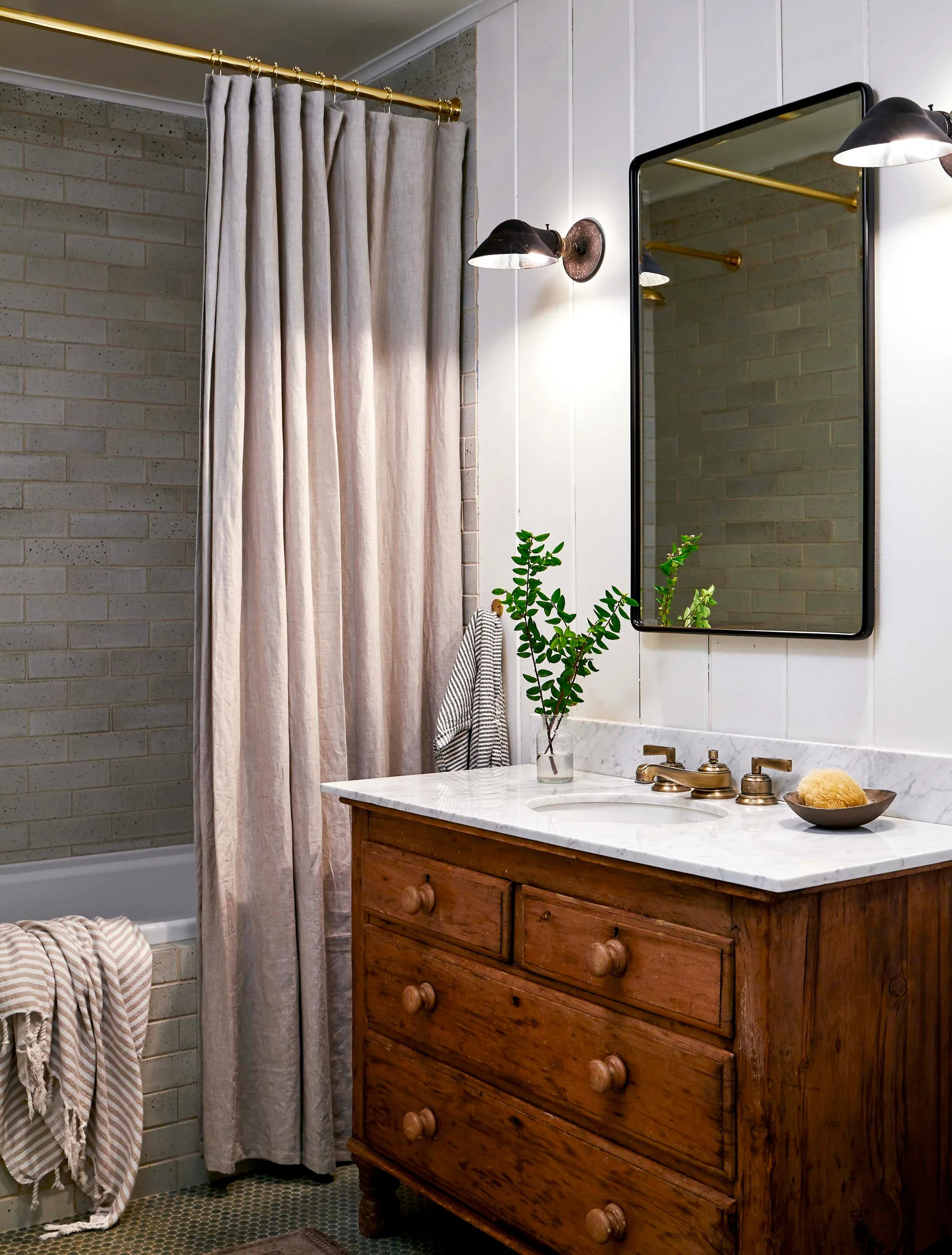 A rustic wooden bathroom vanity with a marble top sits next to a shower with a linen curtain and a brass rod.