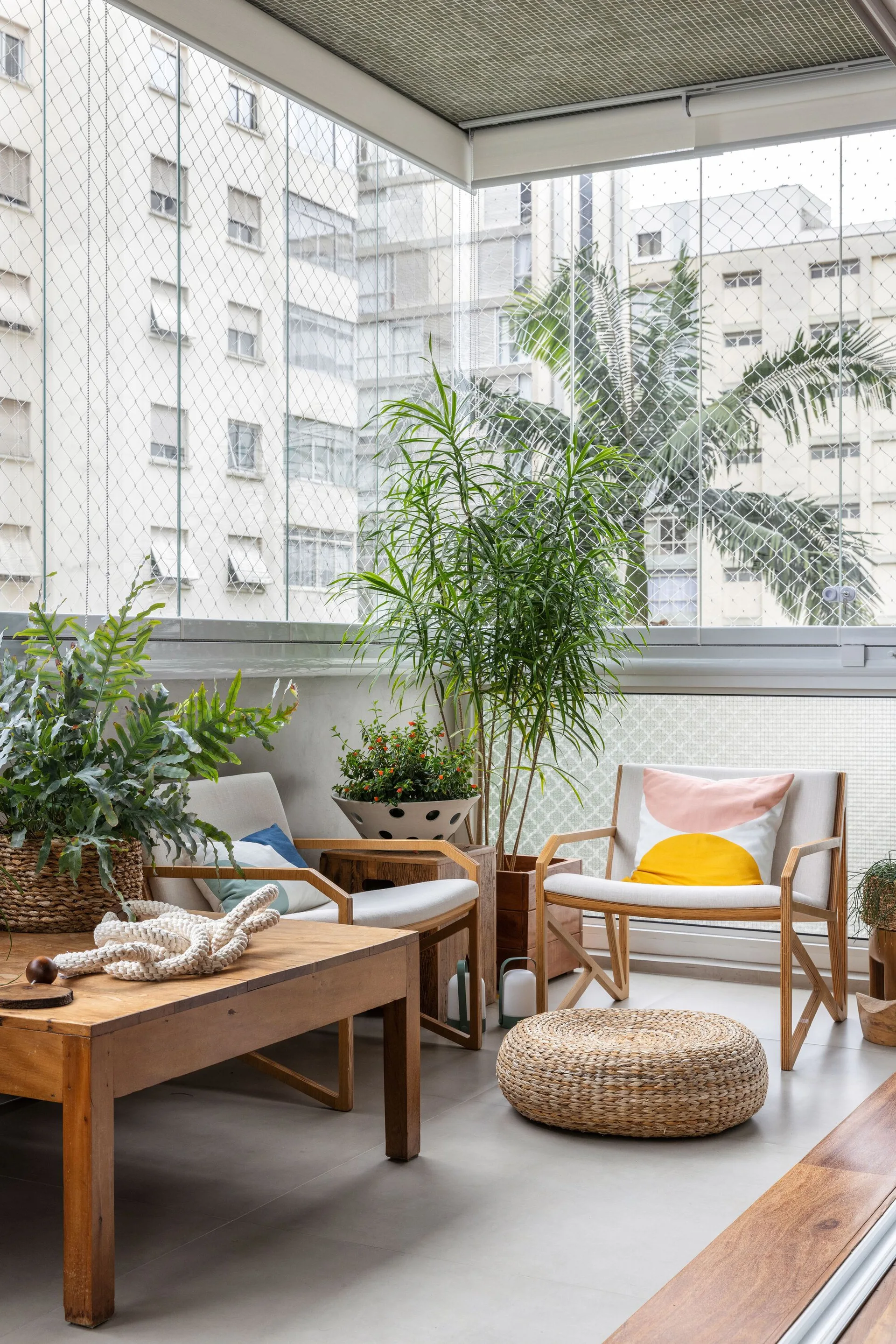 An enclosed urban balcony features wooden armchairs, a woven fiber pouf, a low wood coffee table, and lush potted plants overlooking city buildings.