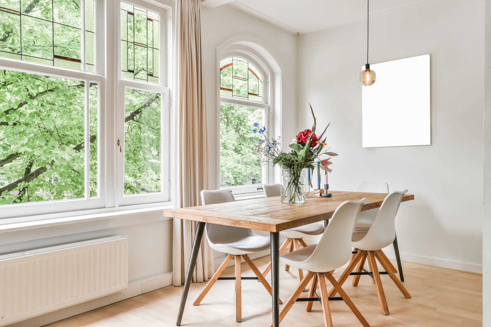 A rustic wood dining table with white chairs stands next to large arched windows with decorative leaded glass panels.