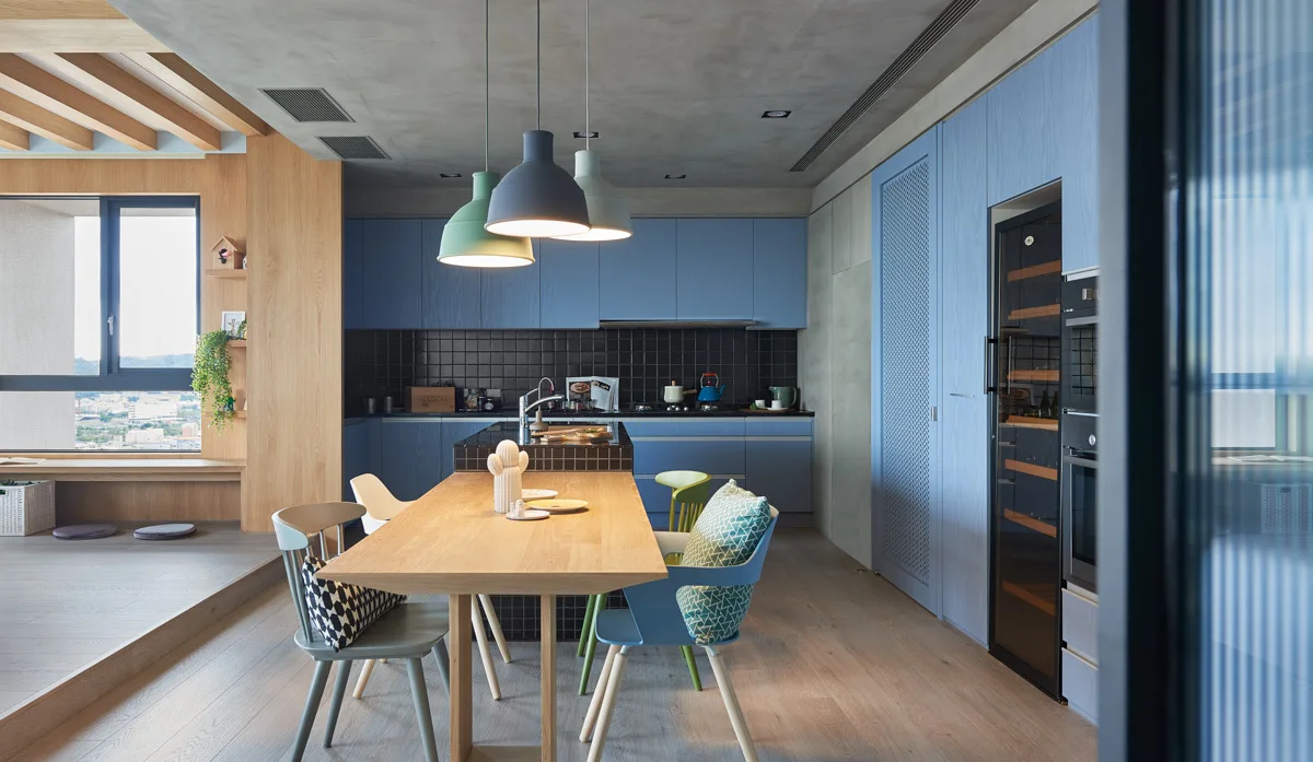 A kitchen with blue cabinetry, a black grid tile island, and an attached light wood dining table with multicolored chairs.