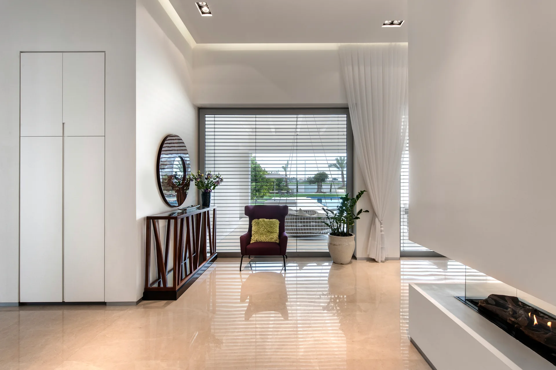 A modern foyer with polished beige floors, a plum armchair by a large window, a wooden console table with a round mirror, and a built-in fireplace.
