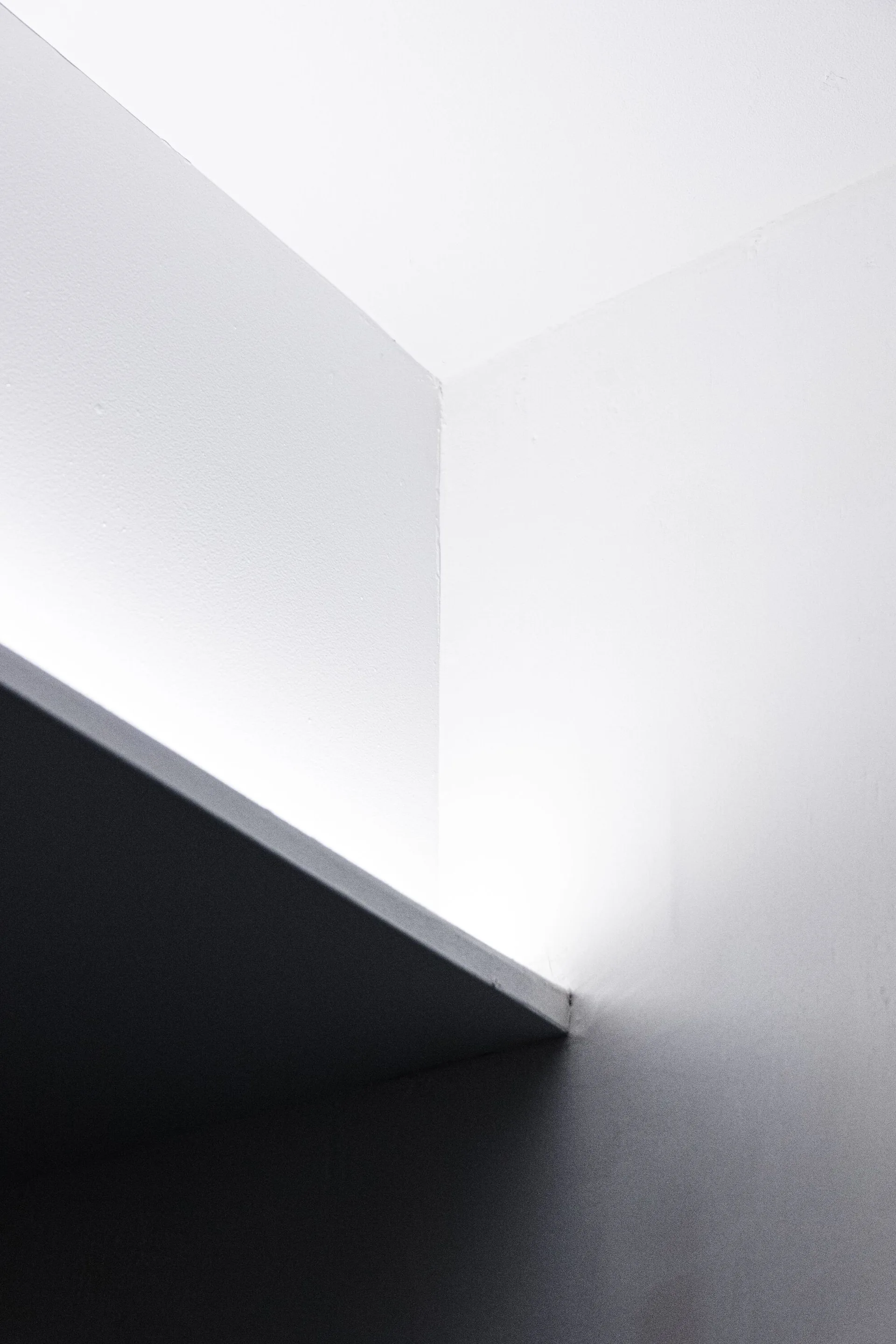 A close-up view of a minimalist white plaster wall corner featuring a horizontal ledge with bright indirect uplighting creating a sharp shadow.