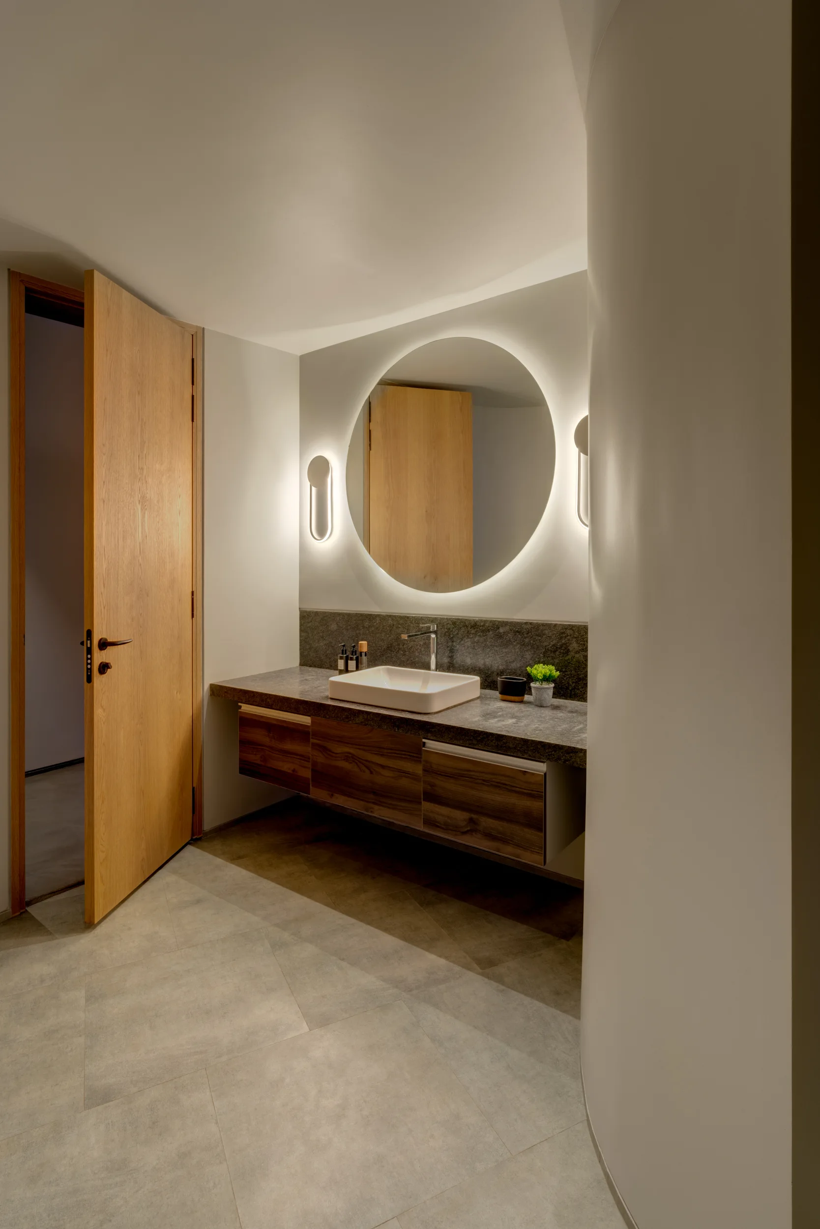 A modern bathroom features a floating wooden vanity with a stone countertop, a white vessel sink, and a large circular backlit mirror.