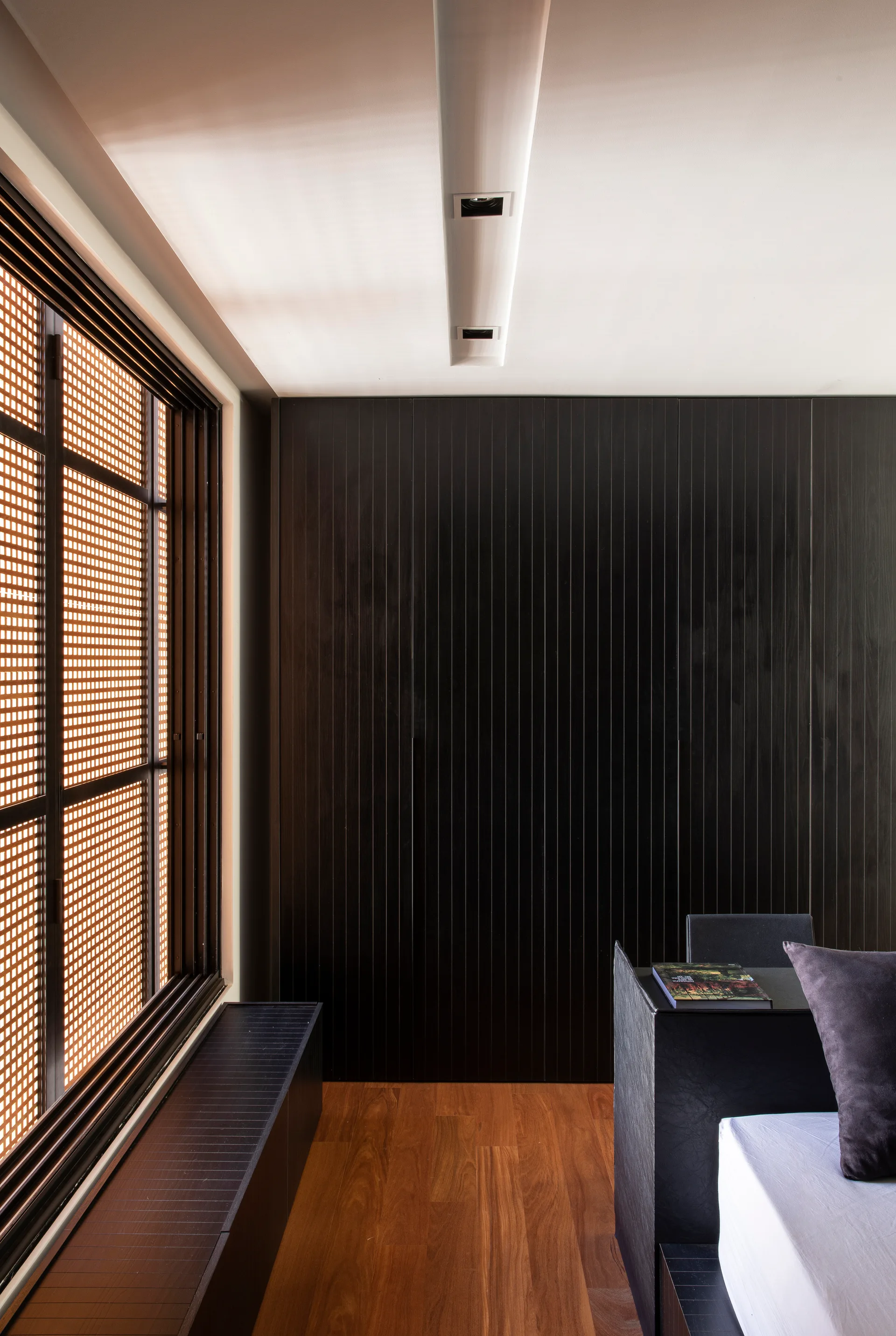 A minimalist bedroom featuring a black vertical slatted wall, a wooden grid window screen, warm wood plank flooring, and a low dark bench.