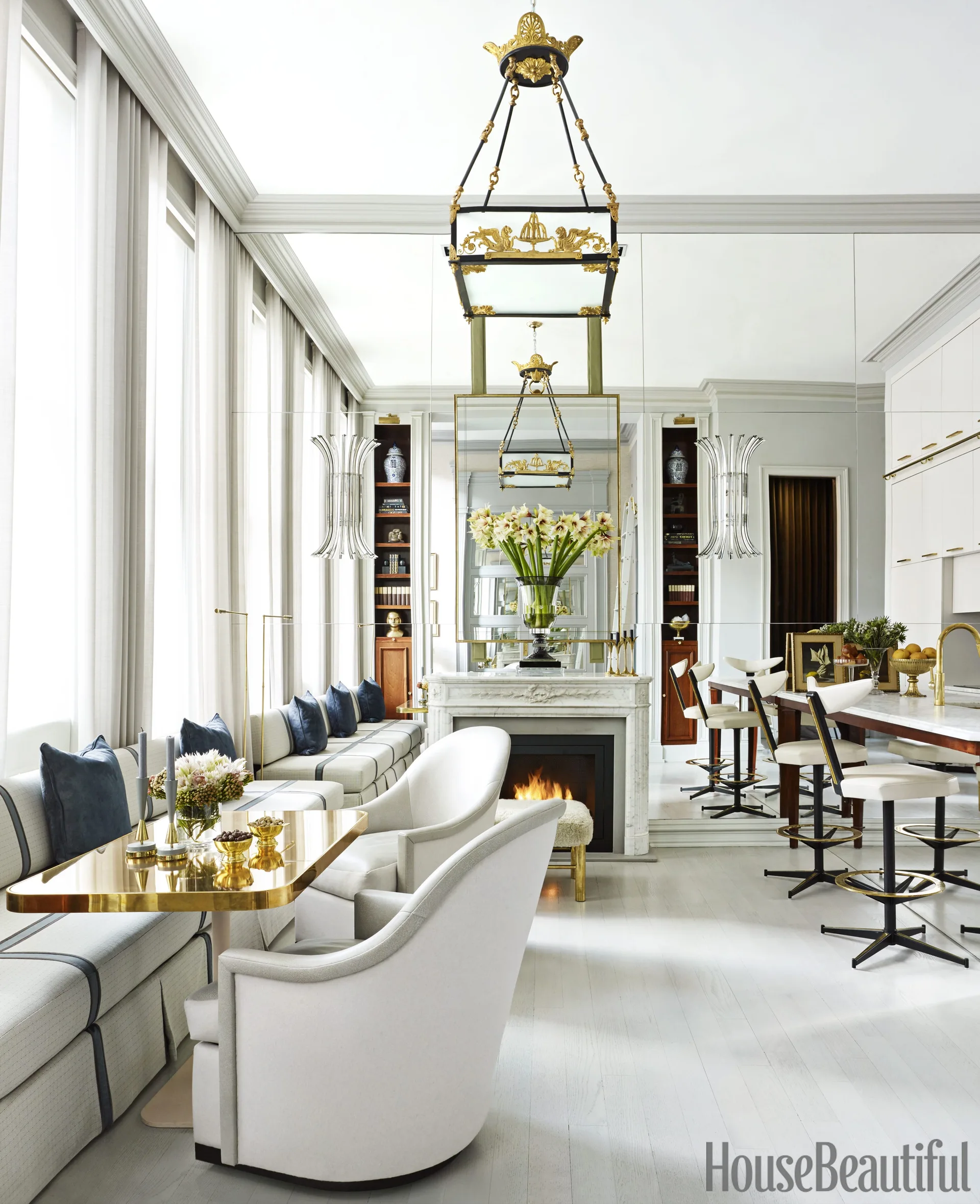 A symmetrical living room features a marble fireplace, a mirrored wall, an upholstered banquette, and a gold dining table with white armchairs.
