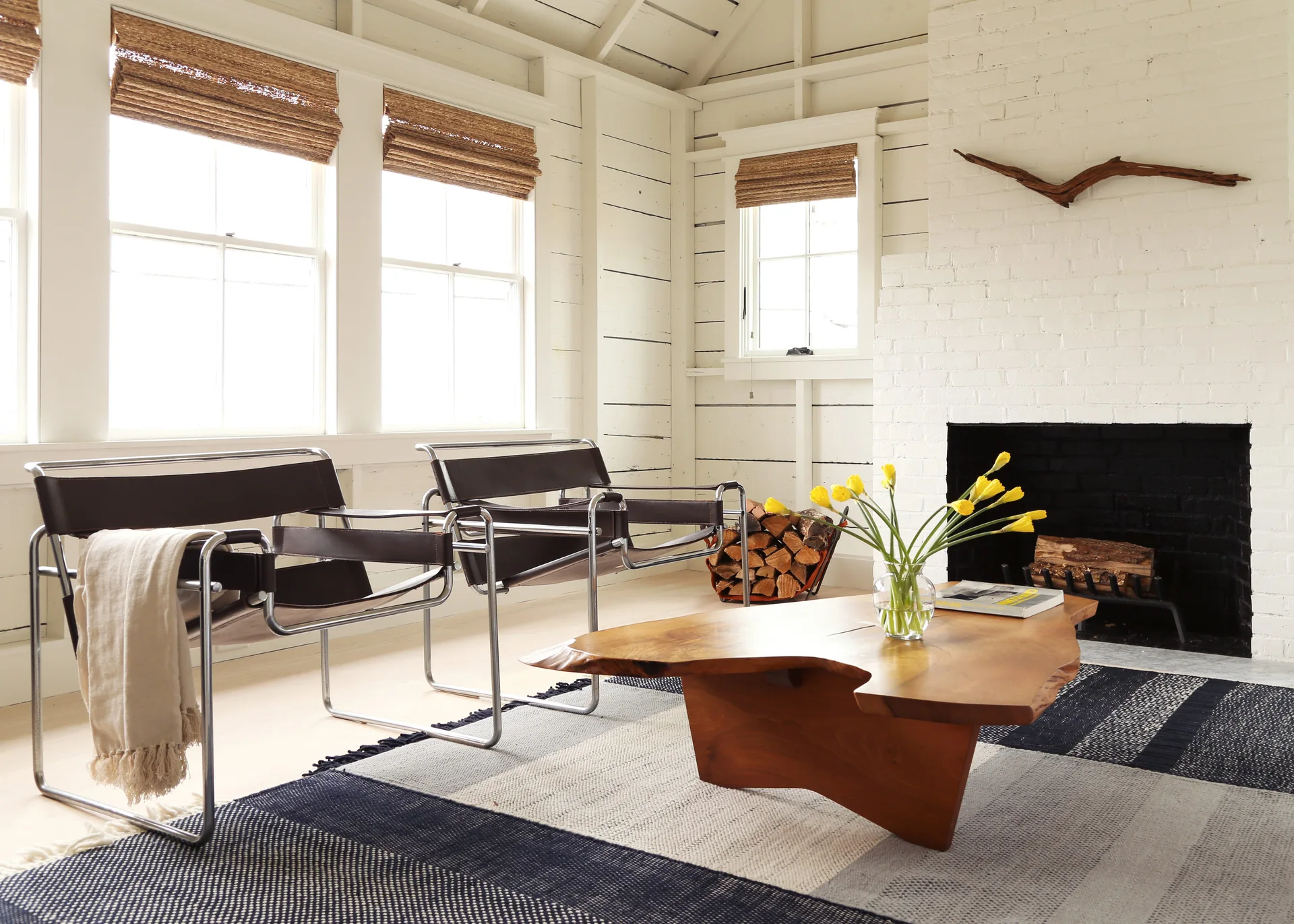 Two brown leather and chrome armchairs face a live-edge wooden coffee table in a bright white living room featuring a painted brick fireplace.