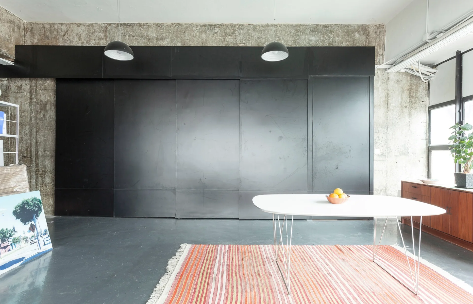 A white oval dining table on a striped rug sits in front of large matte black sliding panels and exposed concrete walls.
