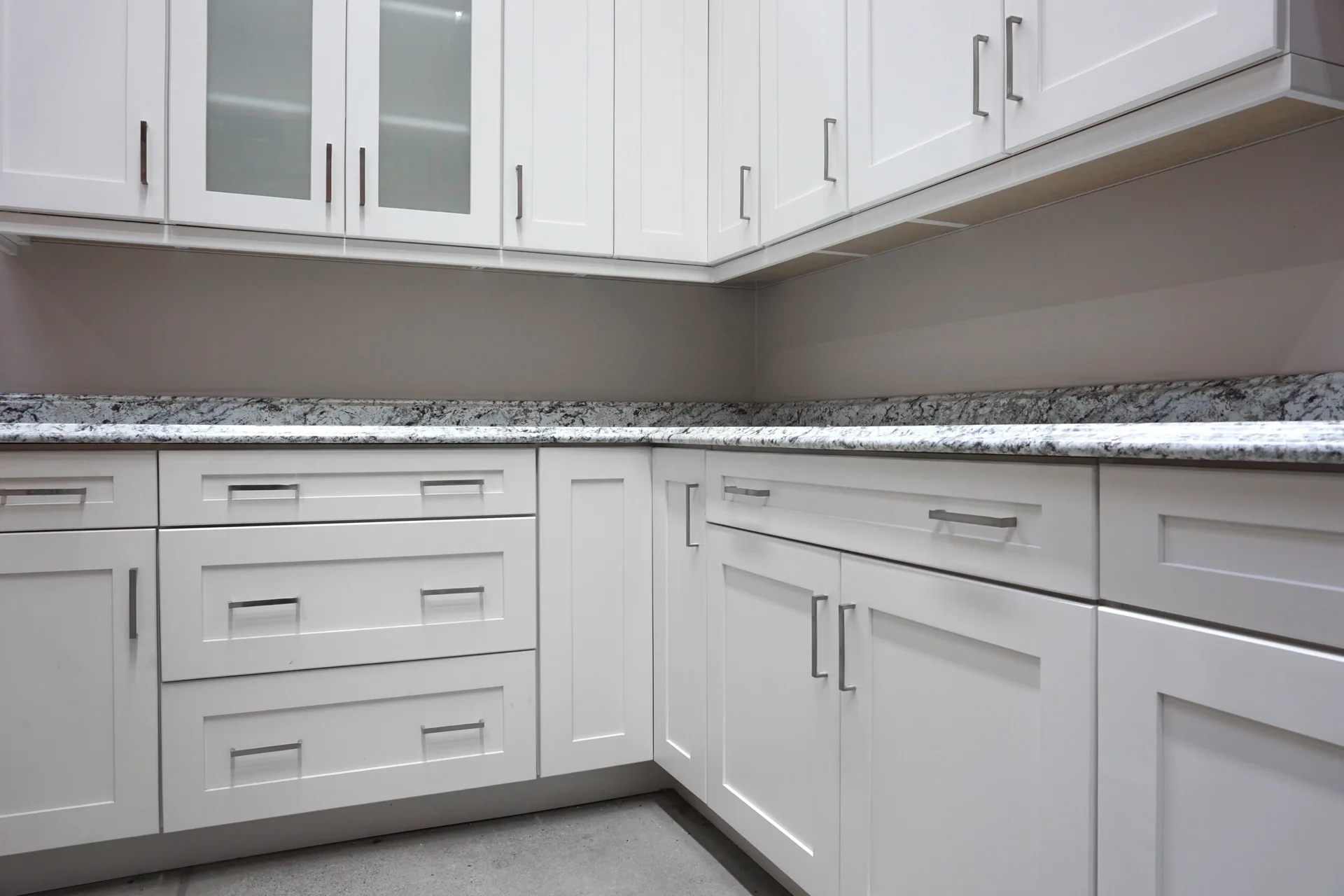 White shaker-style kitchen cabinets with sleek metal drawer pulls are installed below a speckled granite countertop in a corner layout.