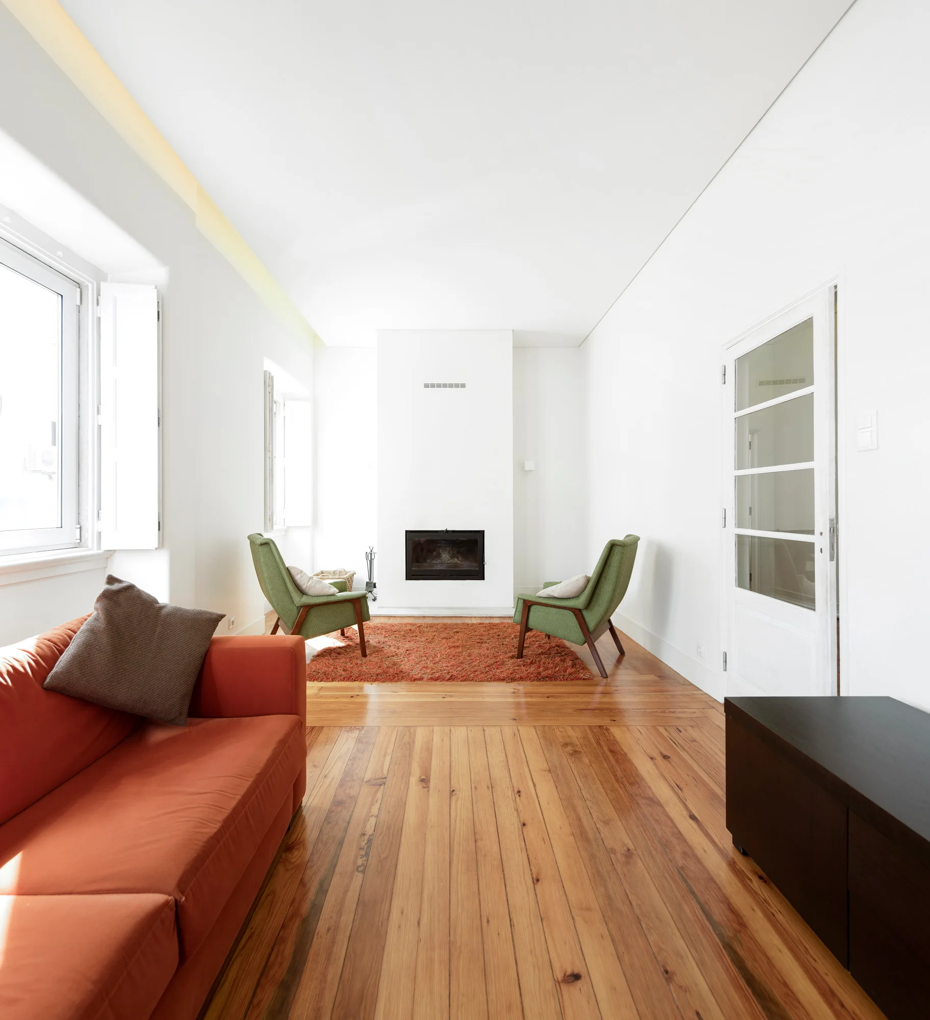 A long white living room features an orange sofa, two mid-century green armchairs, a central fireplace, and polished wood floors.