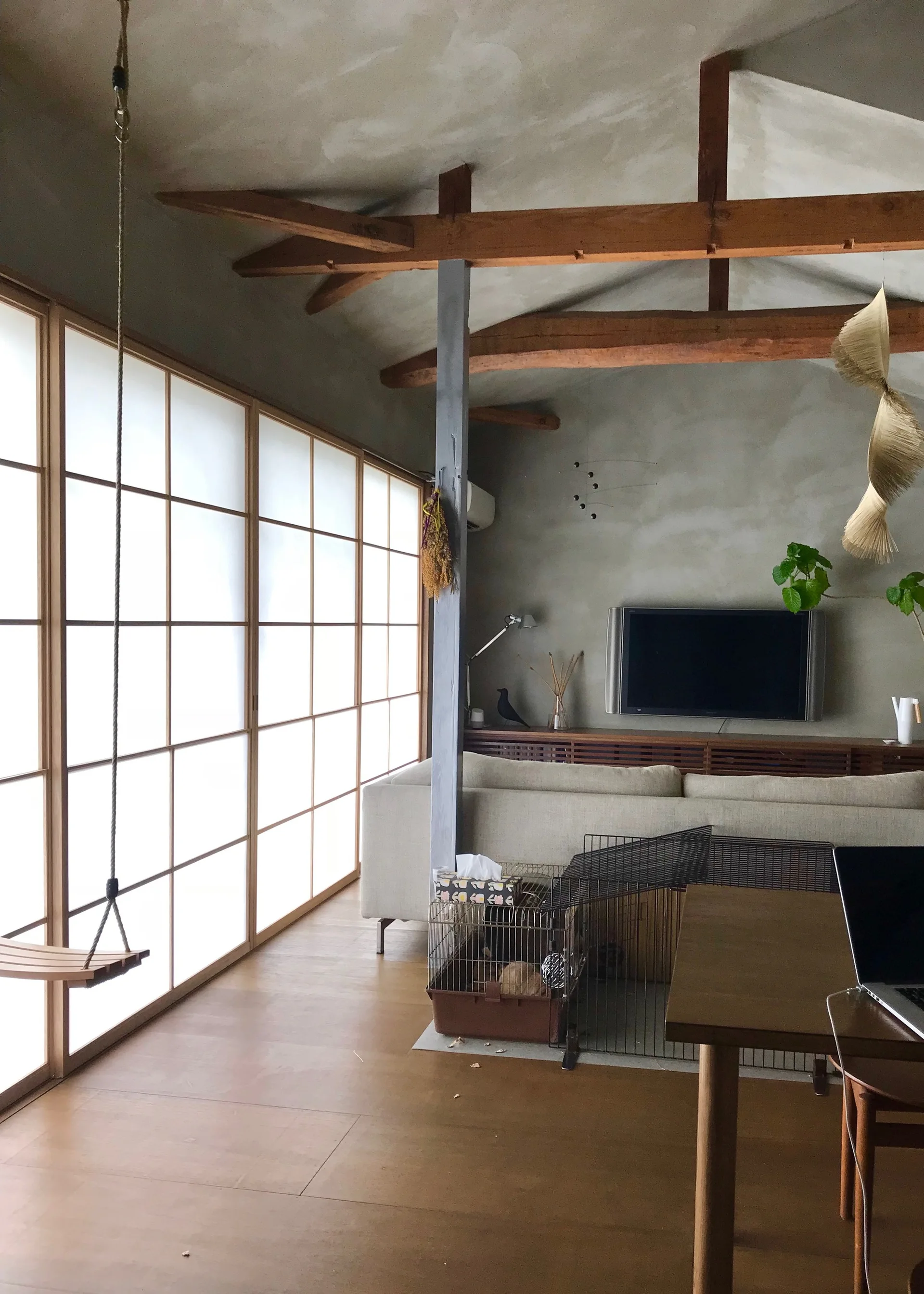 A minimalist living room features large shoji sliding screens, exposed rustic wooden ceiling beams, a light beige sofa, and a hanging wooden swing.