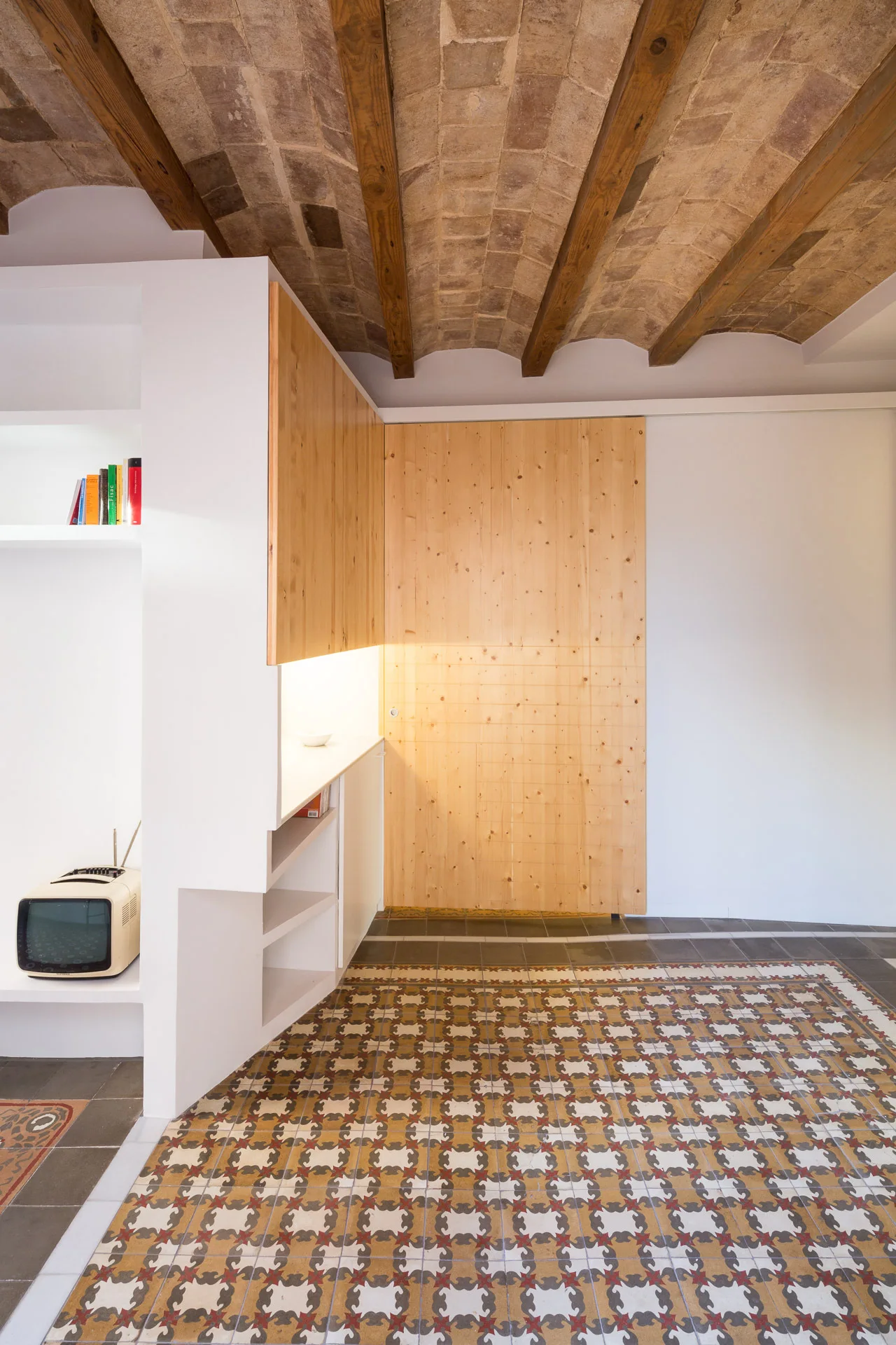 An interior space features a vaulted brick ceiling with timber beams, custom minimalist wood cabinetry, and patterned geometric floor tiles.