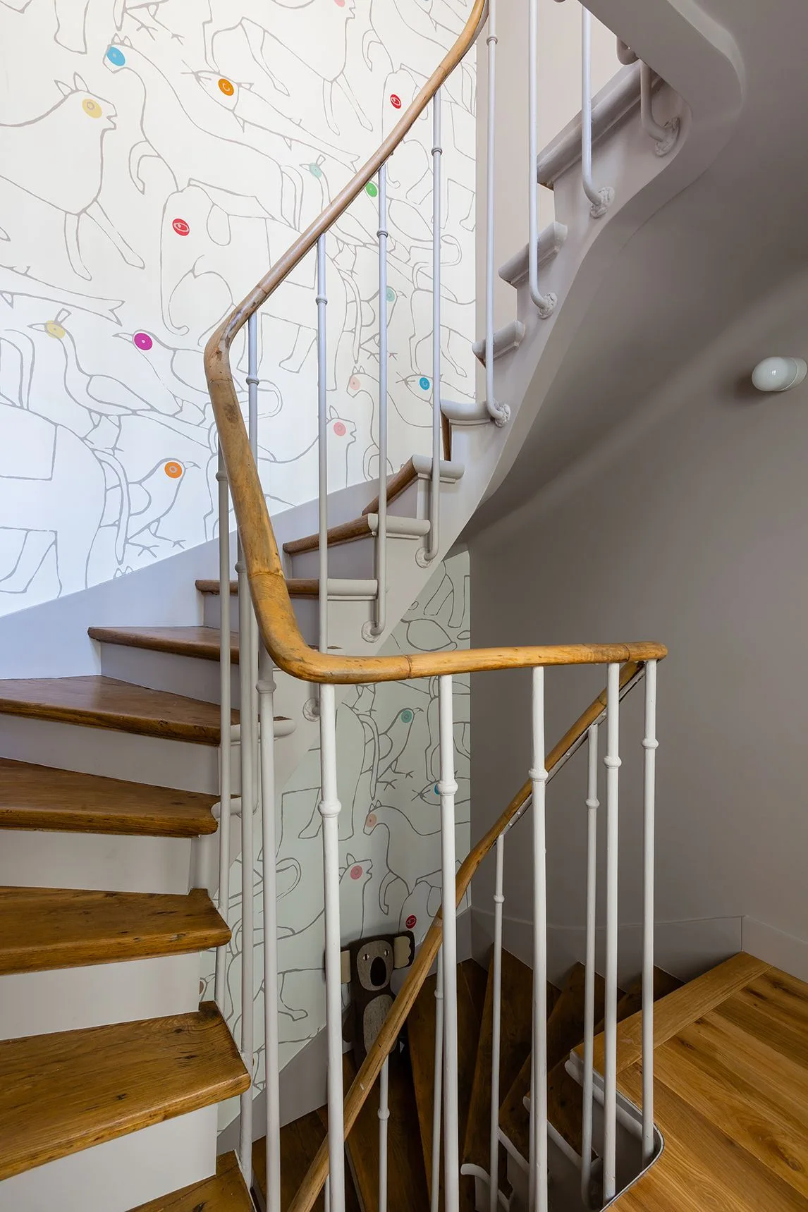 A curved wooden staircase features white metal balusters, a natural wood handrail, and whimsical animal-patterned wallpaper.
