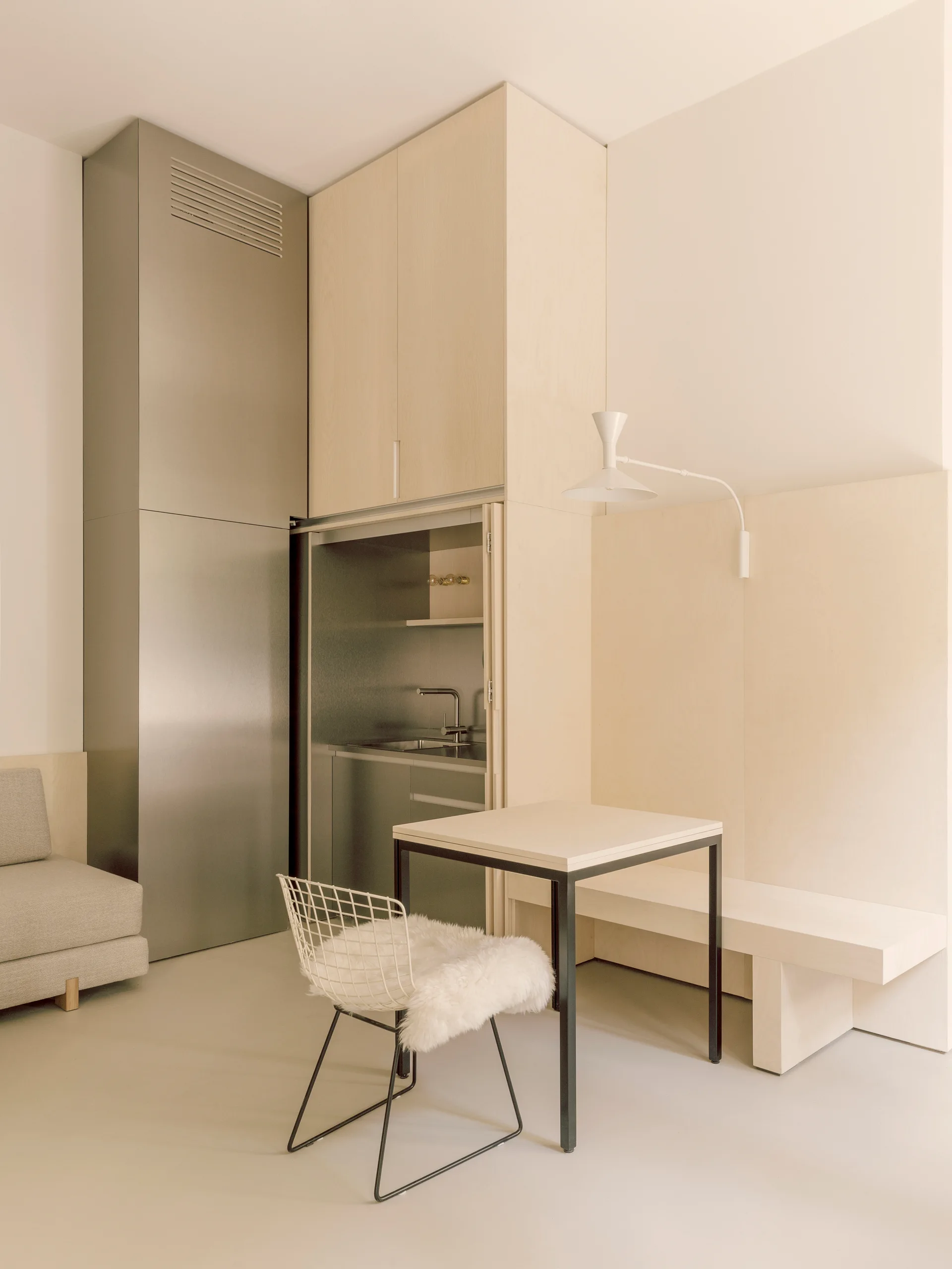 A minimalist studio features a hidden kitchenette built into light wood cabinetry next to a small dining table, wire chair, and a grey sofa.