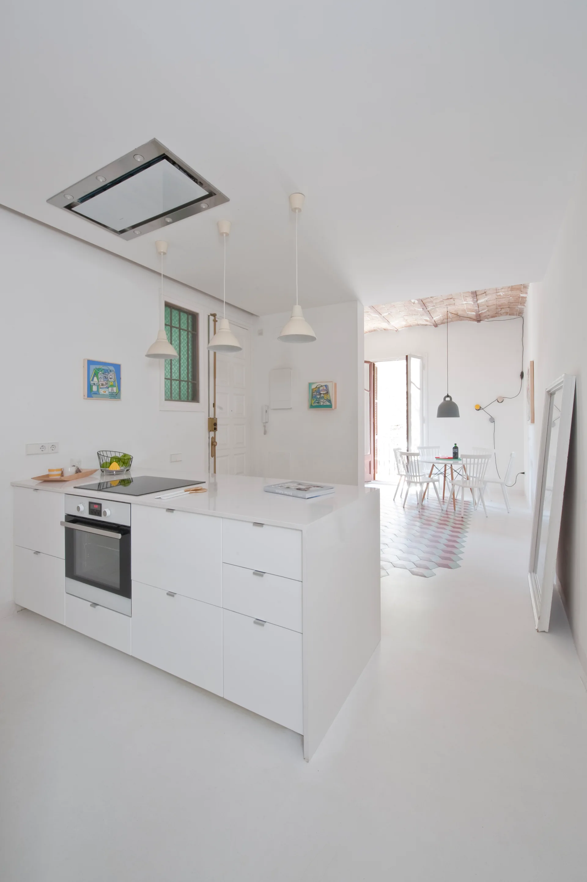 A minimalist white kitchen island with an oven is shown next to a dining area featuring hexagonal floor tiles and an exposed brick vaulted ceiling.