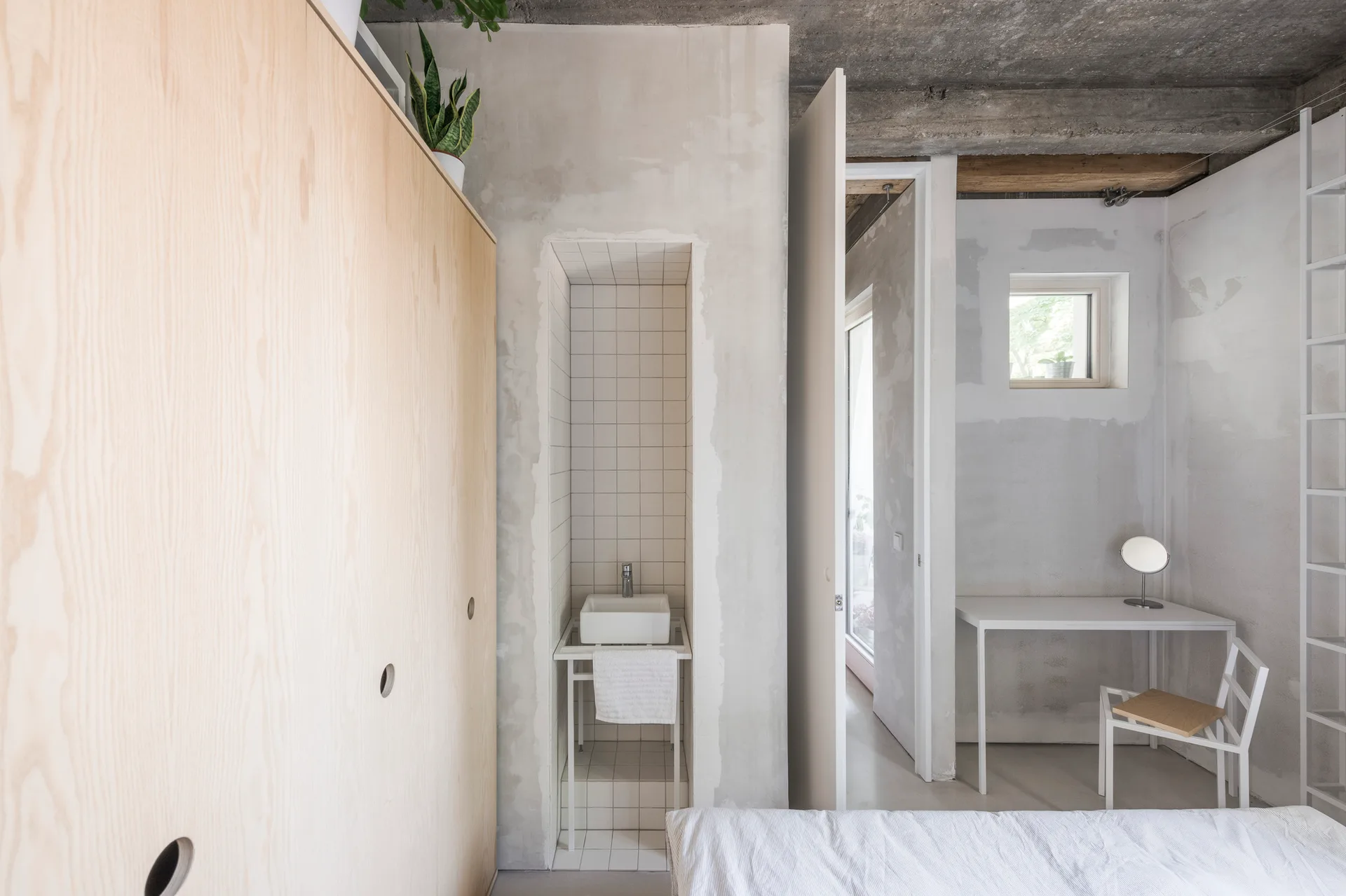 A minimalist bedroom features an exposed concrete ceiling, a large plywood closet, a recessed tile sink alcove, and a simple desk setup.