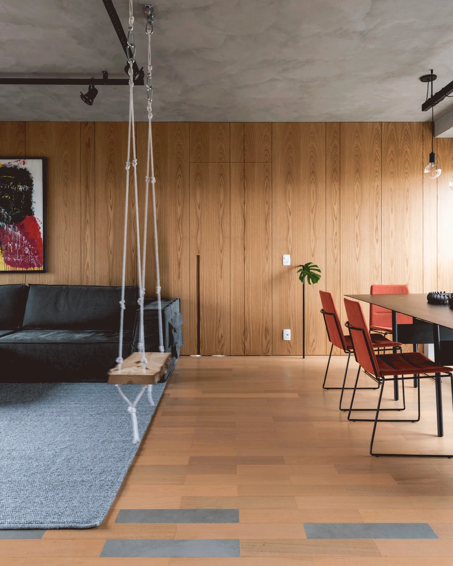 An open living and dining area with a dark sofa, a hanging wooden rope swing, burnt orange dining chairs, and a vertical wood paneled accent wall.