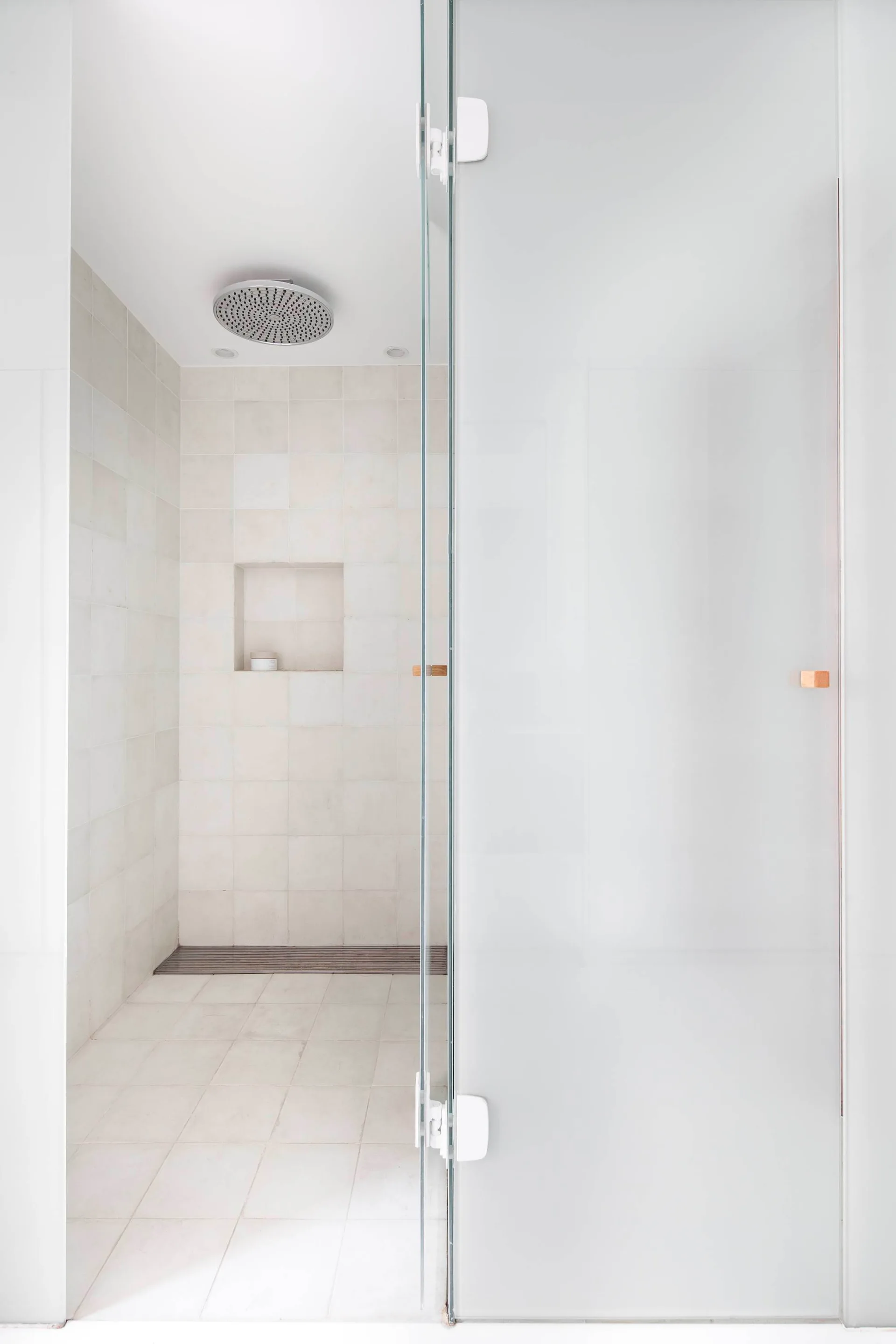 A minimalist walk-in shower features off-white square tiles, a recessed wall niche, a linear drain, and a frosted glass door.