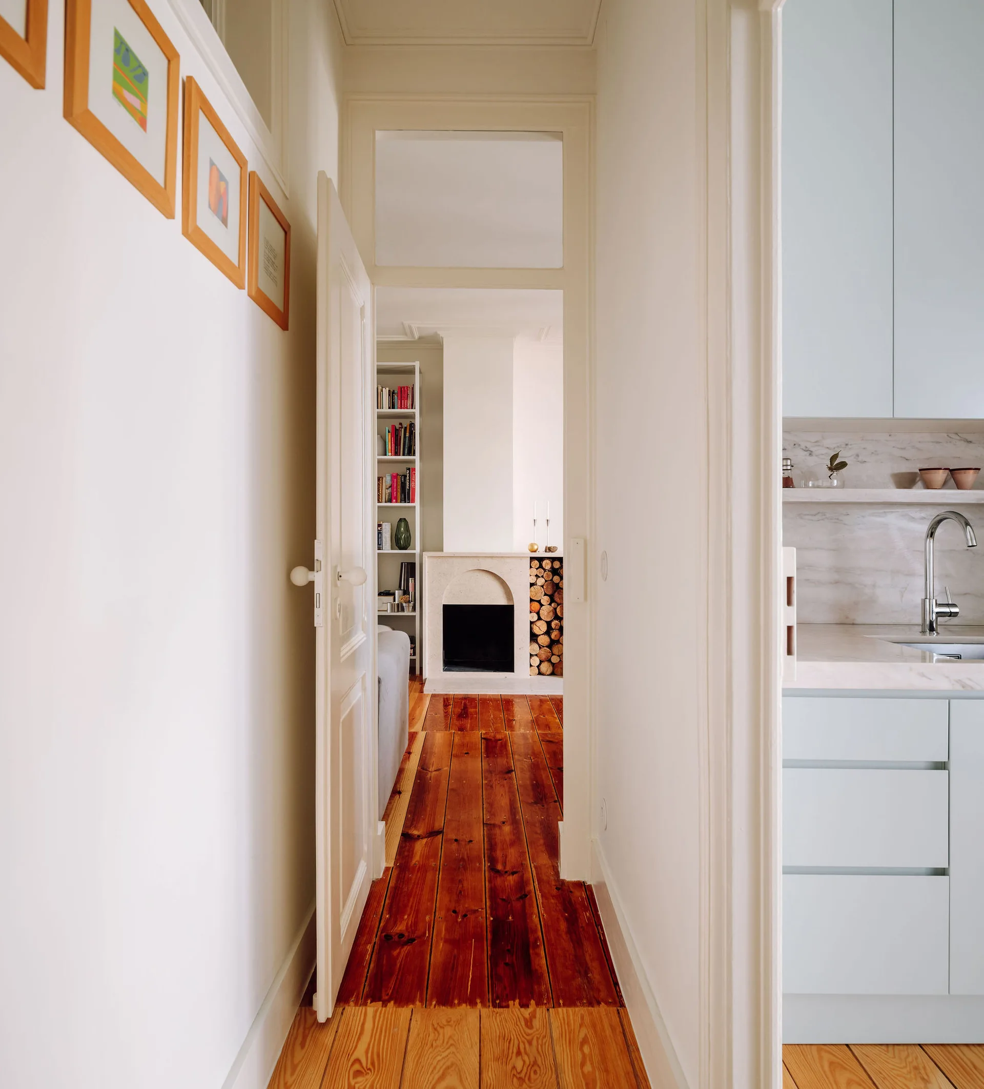 A narrow wood-floored hallway connects a pale blue kitchen on the right to a white living room with a stone fireplace.