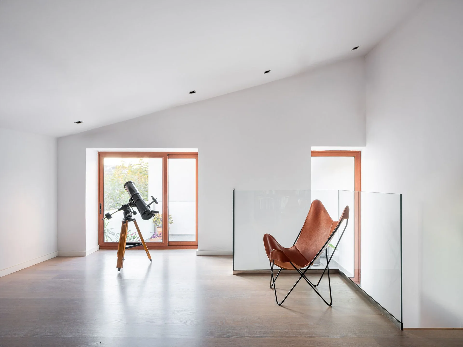 A tan leather butterfly chair sits next to a glass railing on a minimalist landing with white walls, a pitched ceiling, and a telescope near a window.