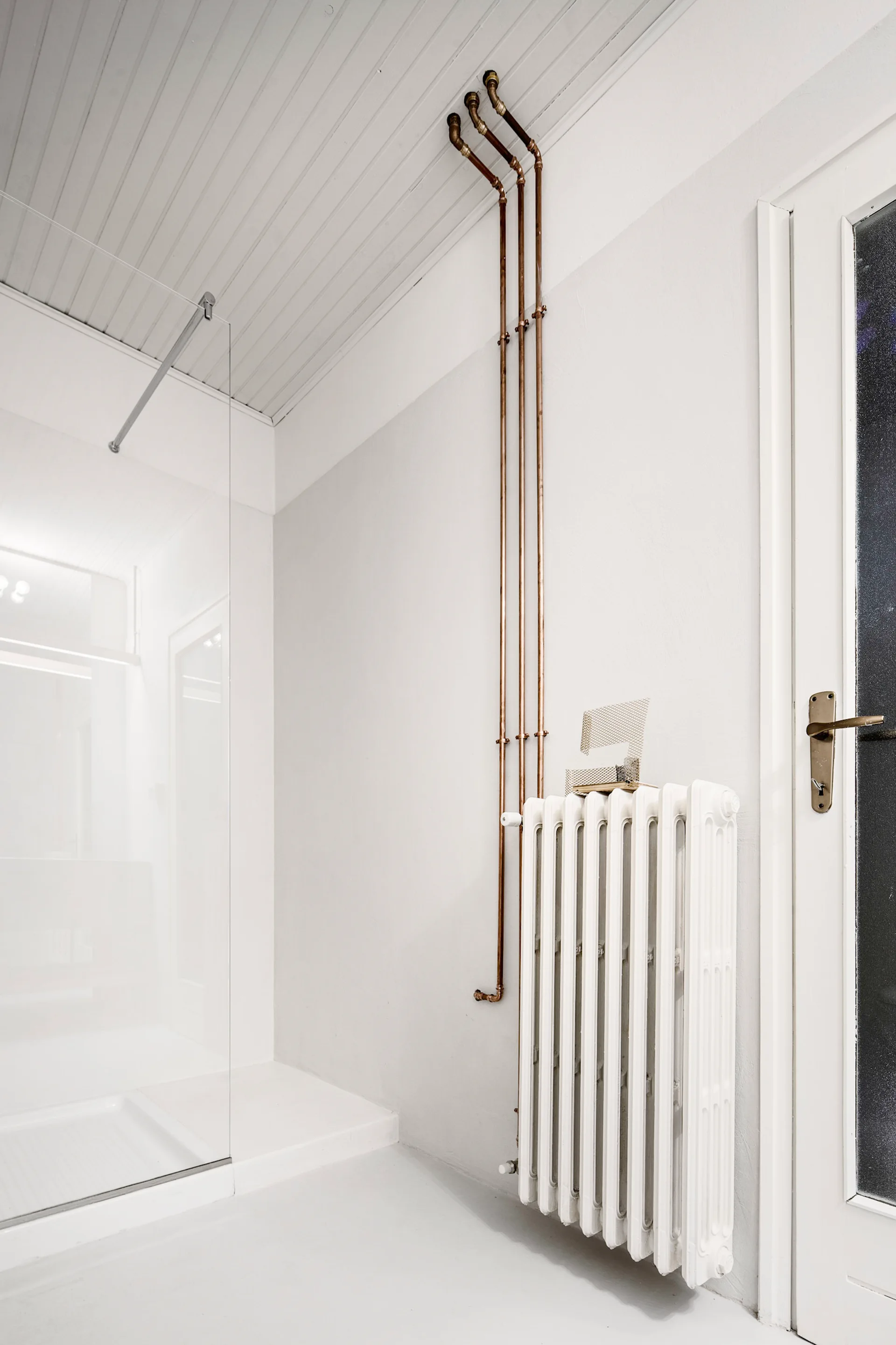 A minimalist white bathroom features vertical exposed copper pipes on a plain wall next to a classic white cast iron radiator.