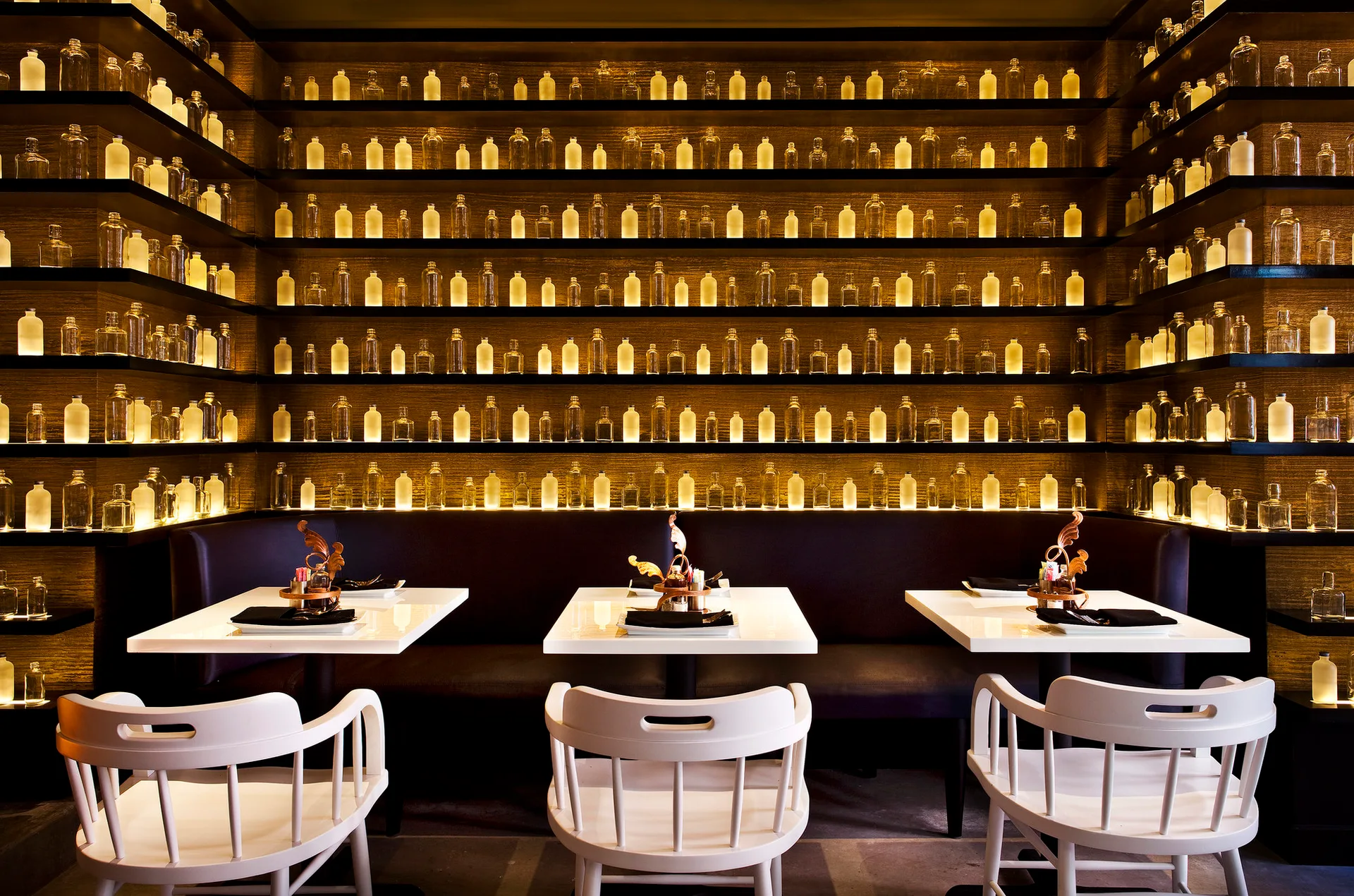 White dining tables and spindle-back chairs sit in front of a dark banquette and walls lined with multiple rows of backlit glass bottles.