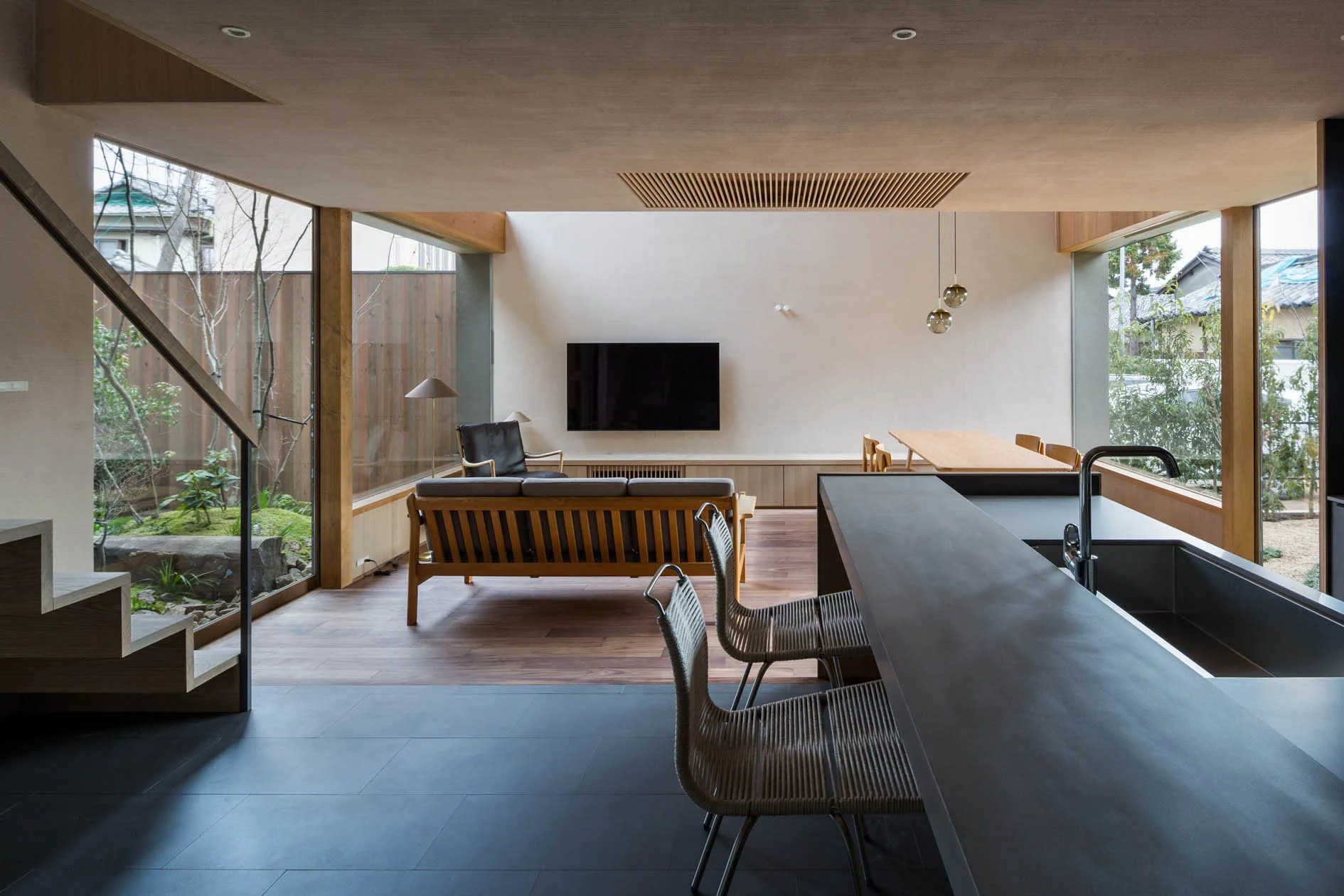 An open-plan living area features a dark kitchen island with woven barstools, wood floors, and floor-to-ceiling windows showing a pocket garden.