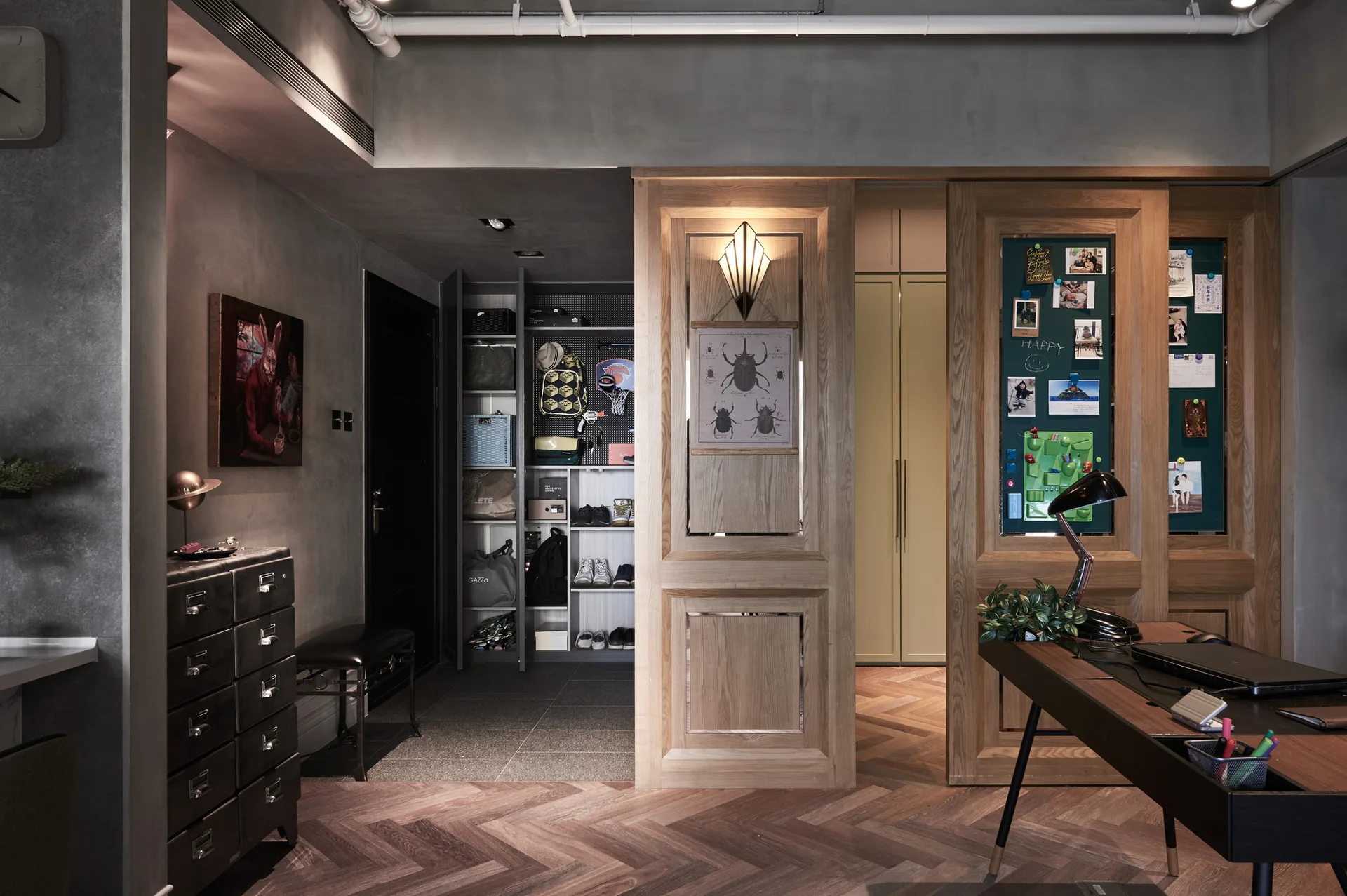 An industrial home office features herringbone wood flooring, grey concrete walls, a dark wood desk, and large wood-paneled sliding divider doors.