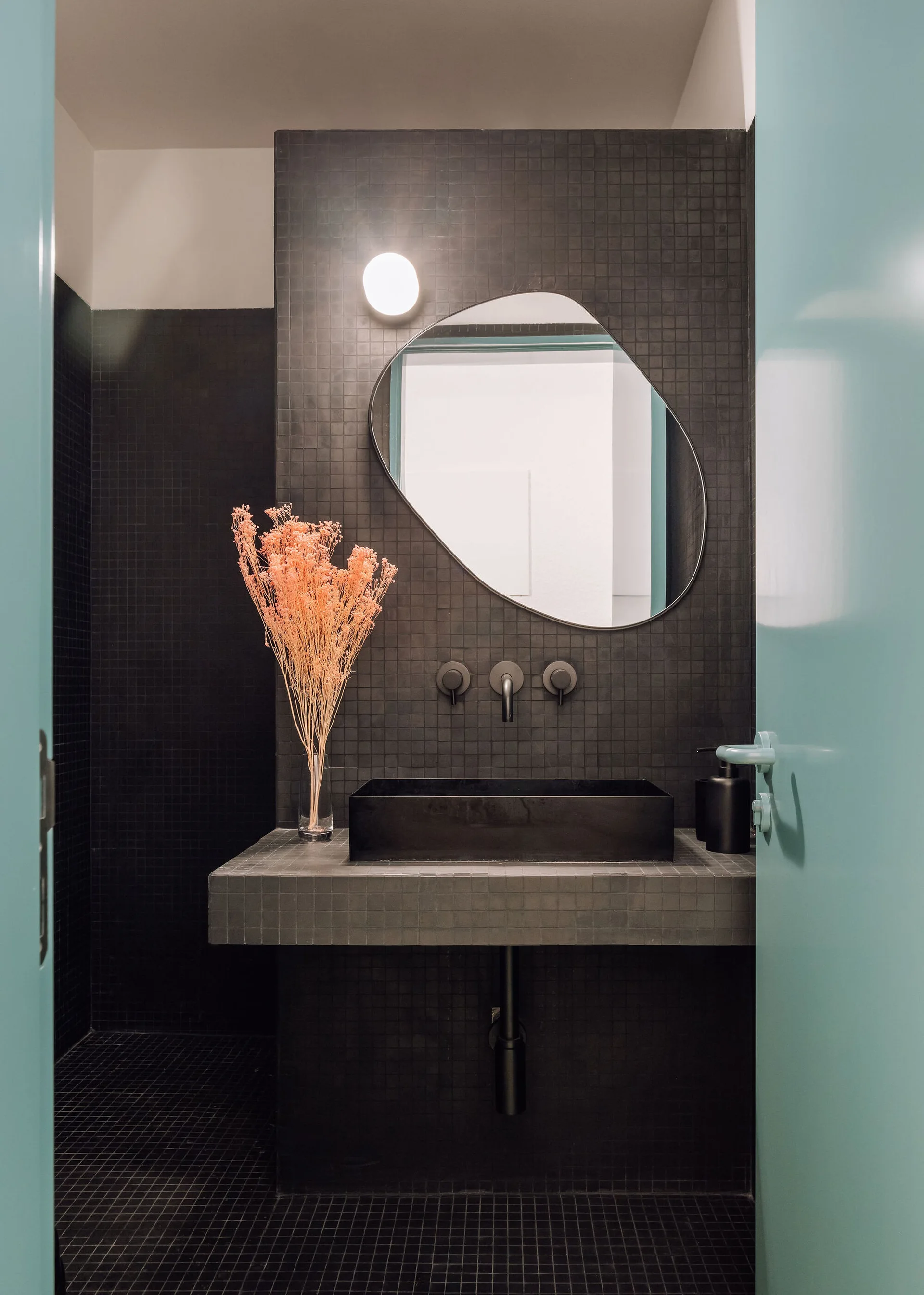 A modern black bathroom features a rectangular sink on a dark mosaic-tiled vanity, an asymmetrical mirror, and a bright teal door frame.